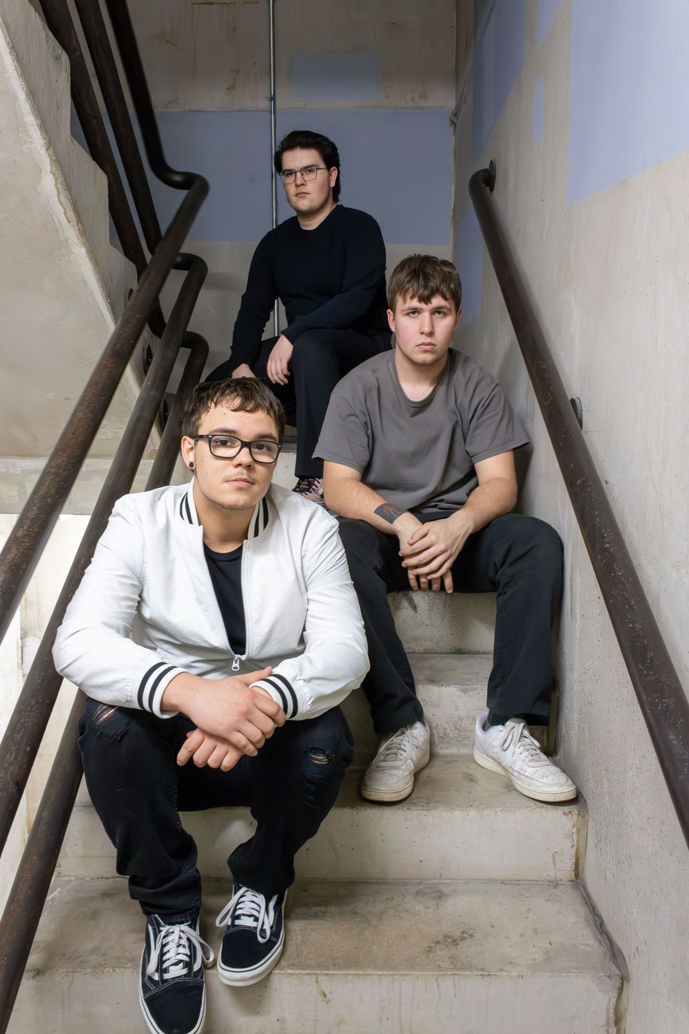 Three musicians sit casually on concrete stairs wearing casual clothing in an indoor setting.