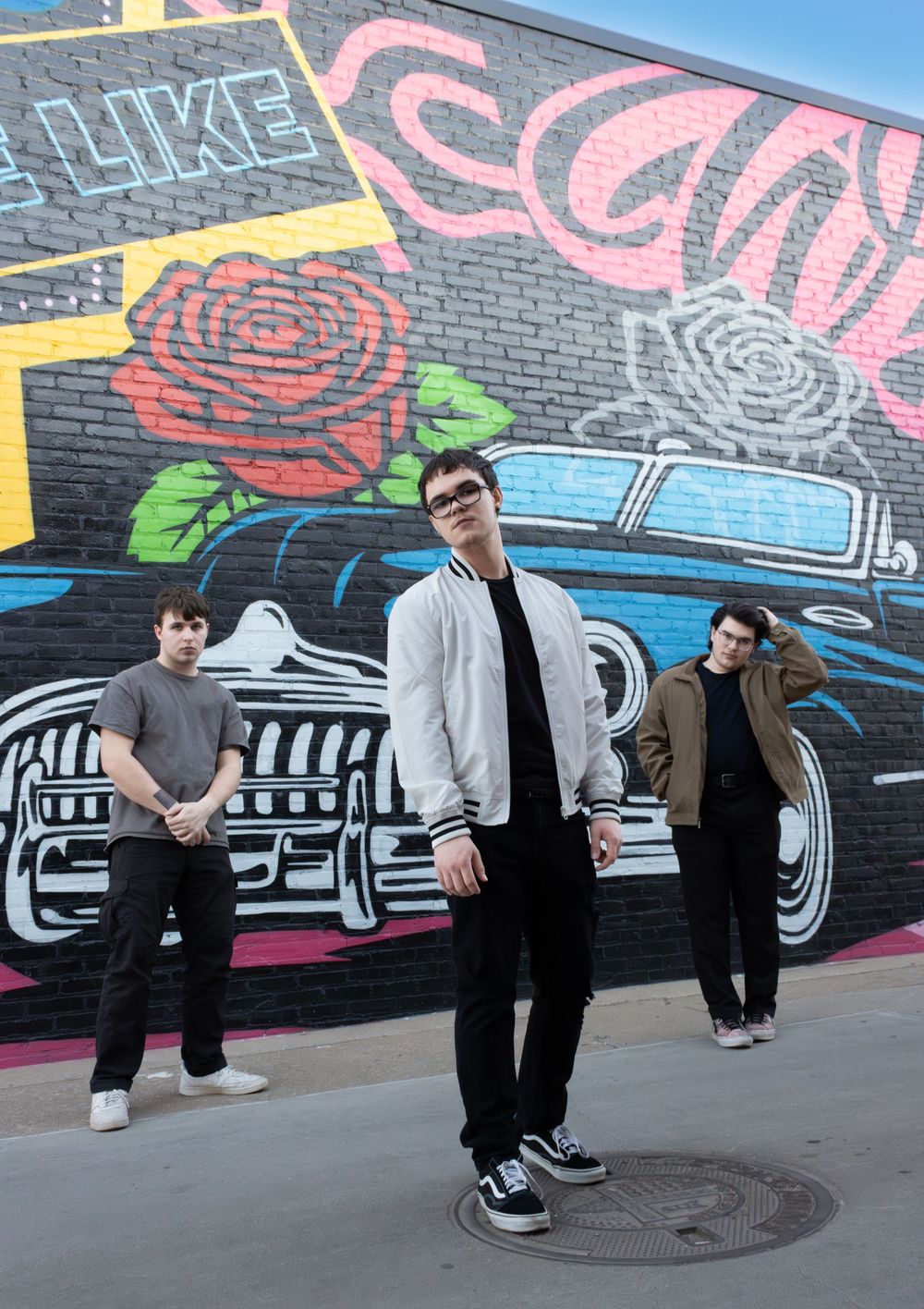 Three people pose in front of a colorful street mural featuring roses and classic cars on a brick wall.