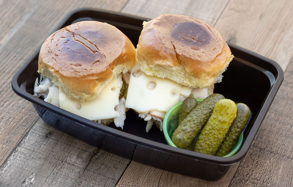 Small sliders sit in takeout containers with dill pickles on rustic wooden table surface.