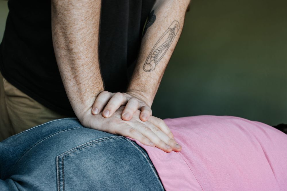 Tattooed hands perform a chiropractic manipulation on a person wearing pink and denim clothing.
