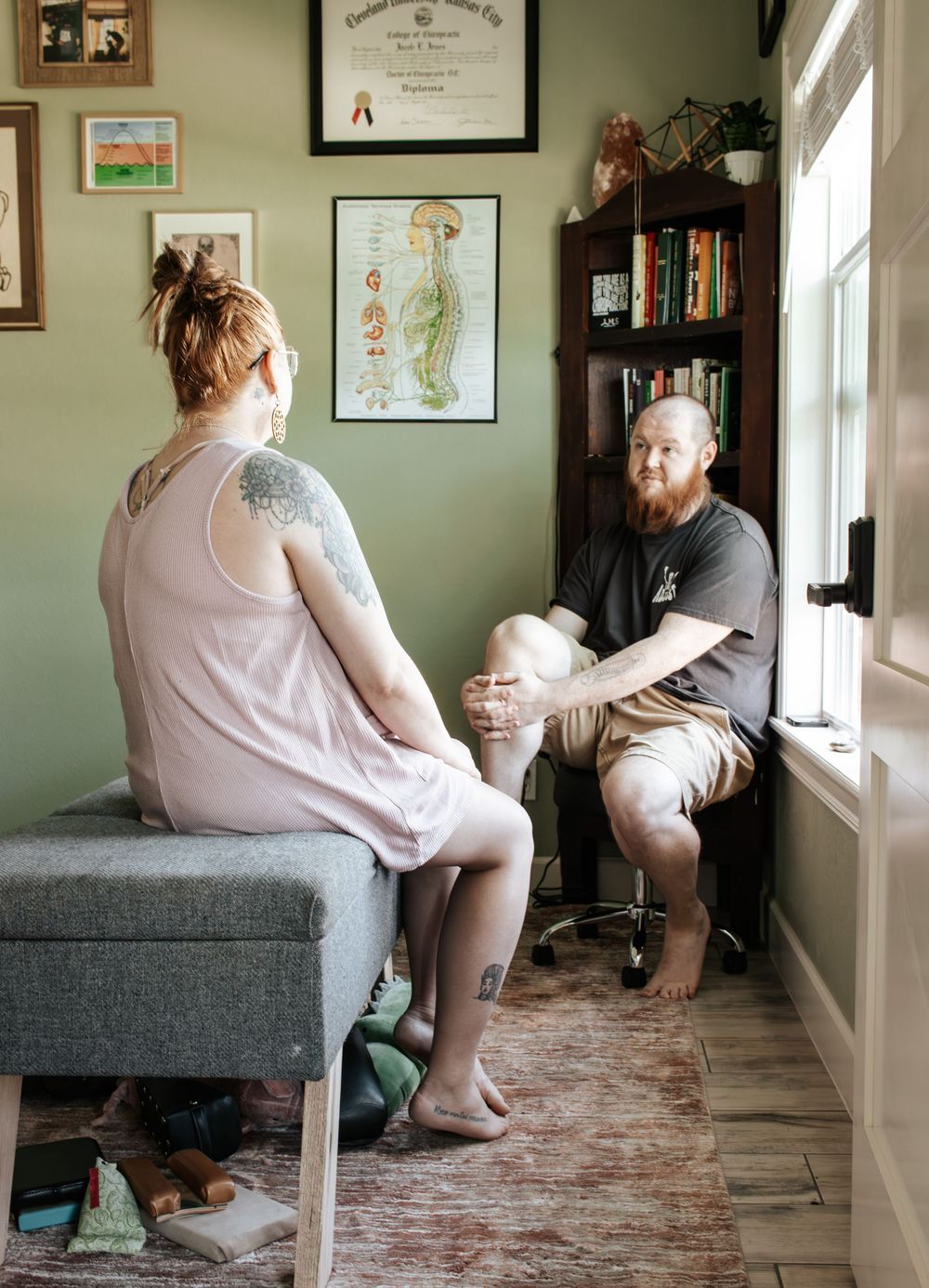 Two people sit in conversation near a window in a cozy home office space with green walls and framed artwork.