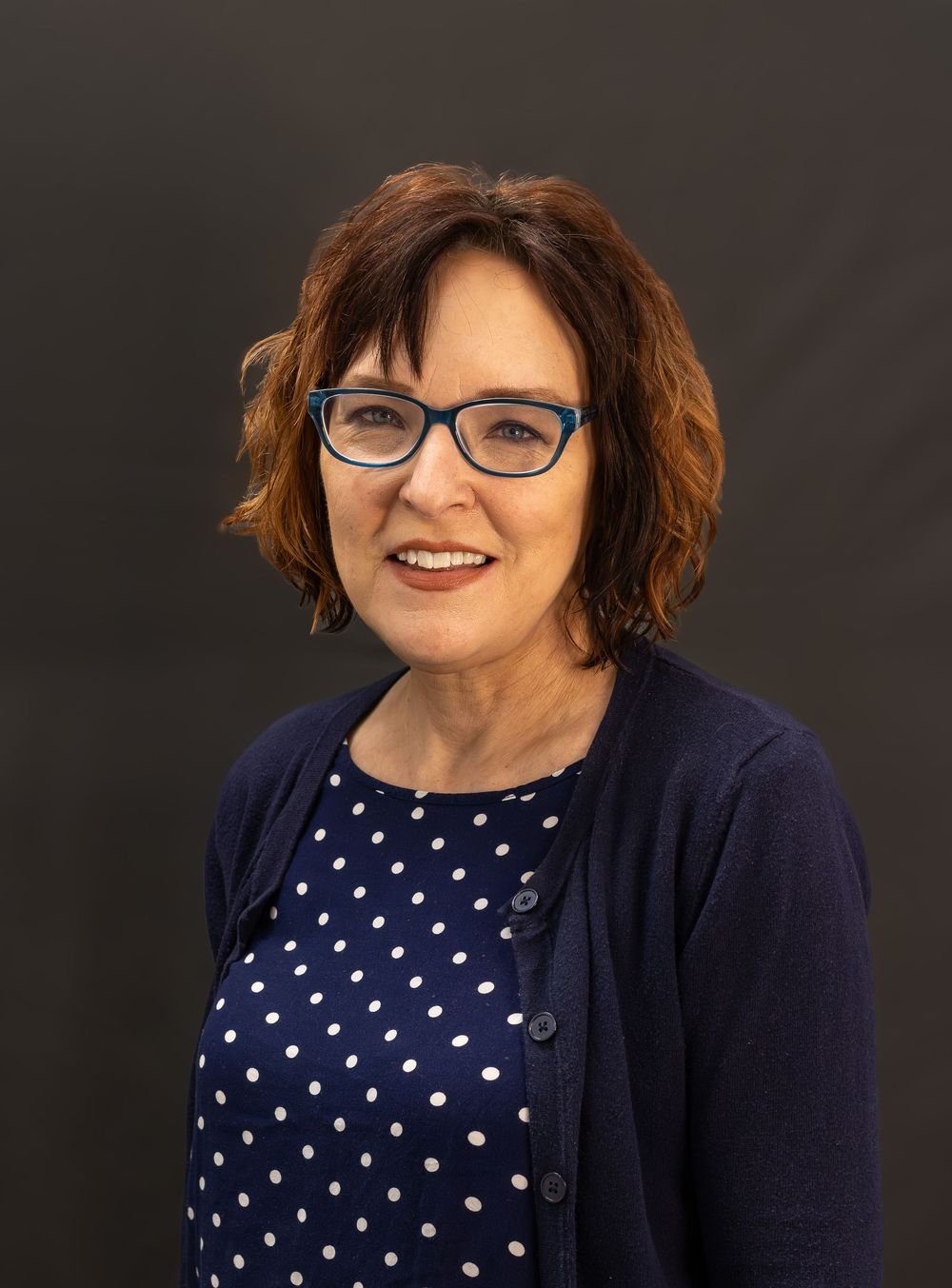 Professional portrait of someone wearing glasses and a navy cardigan over a polka dot blouse against a dark background.