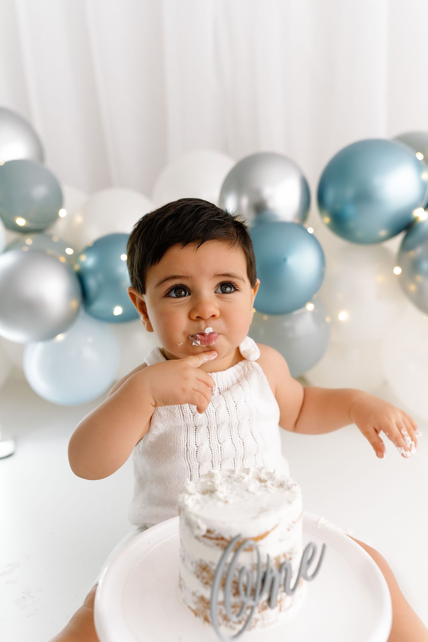 First birthday celebration setup with blue and silver balloons and white smash cake against white backdrop.