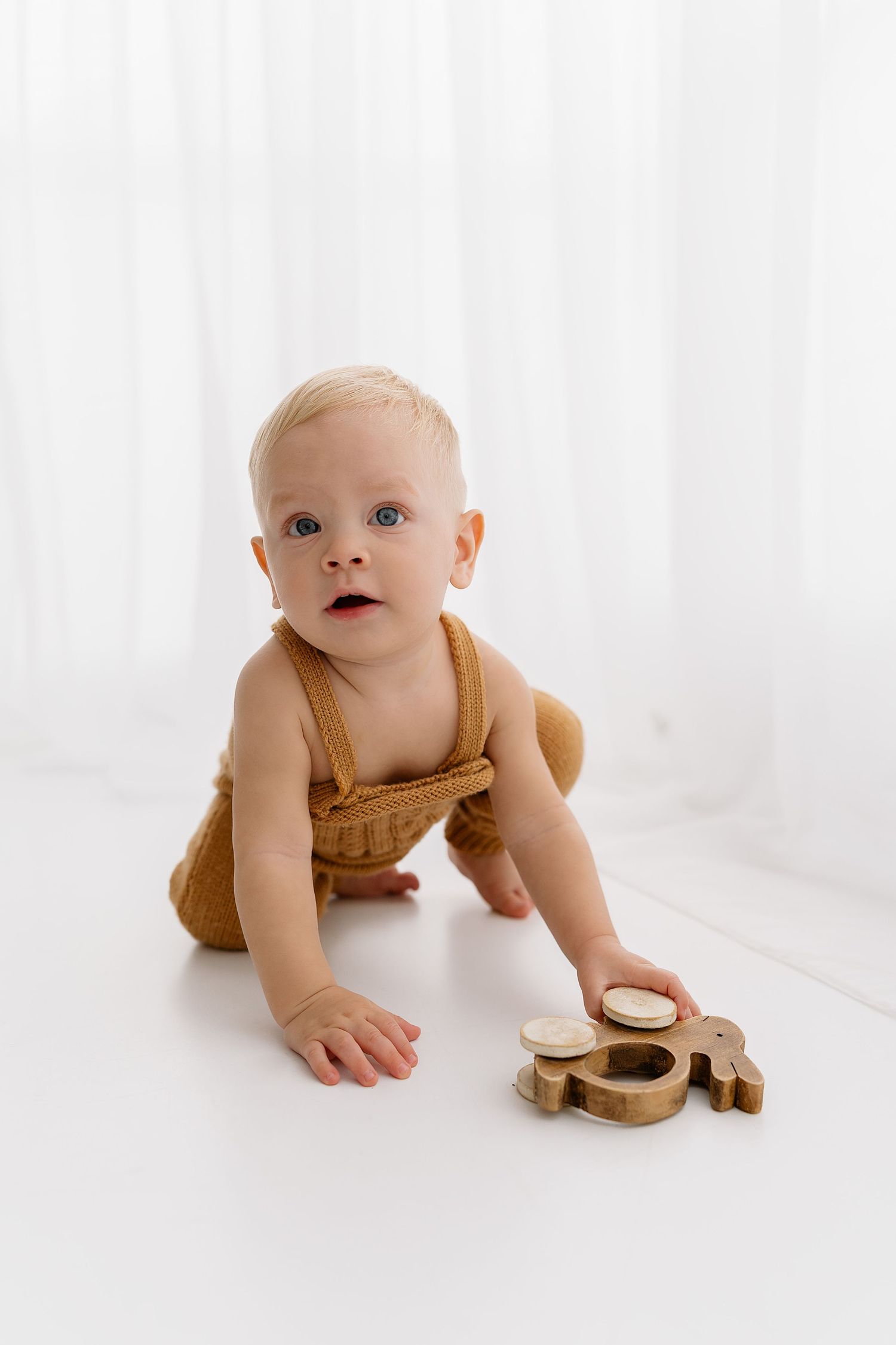 Baby wearing mustard-colored outfit crawls on white floor while playing with wooden toys.