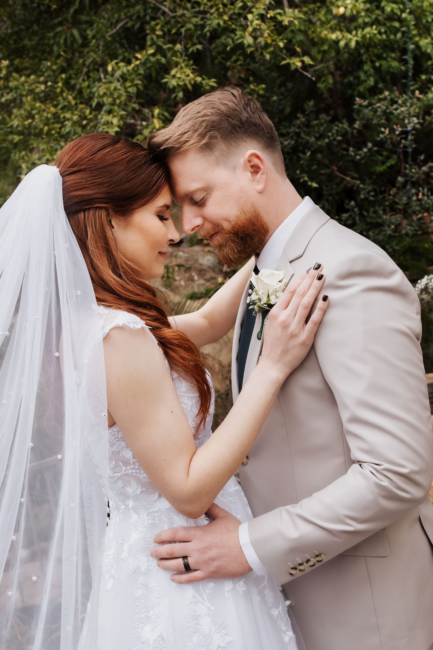 Romantic wedding couple touching foreheads outdoors, bride in white lace gown with veil, groom in beige suit with boutonniere.