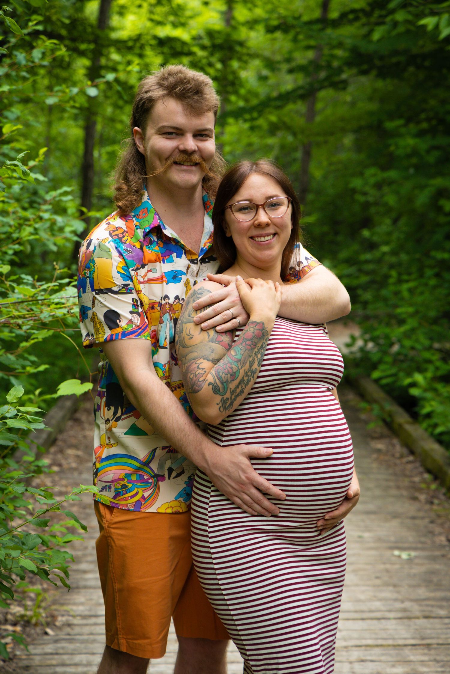 Expecting couple poses on wooden walkway surrounded by lush forest greenery.
