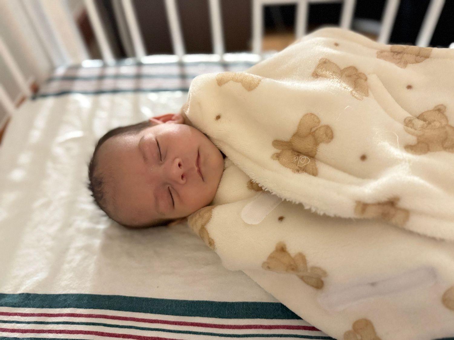 A sleeping baby bundled in a cream colored blanket with teddy bear prints rests on striped bedding.