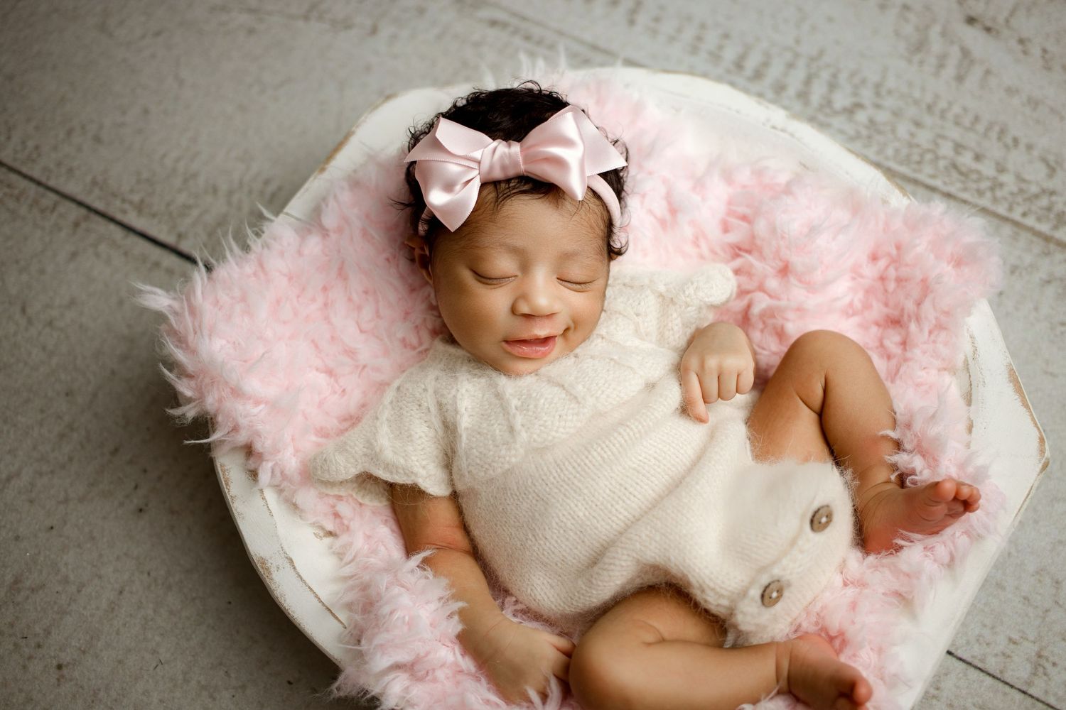 Sleeping newborn wearing white outfit and pink bow rests peacefully on soft pink fur blanket.