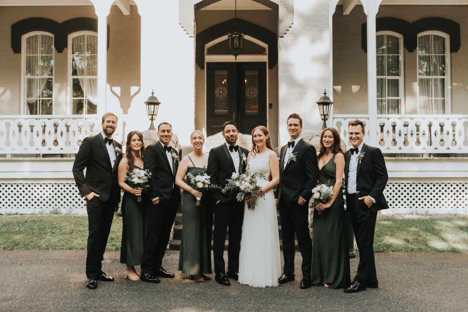 Wedding party in formal attire poses together on steps of elegant white historic mansion with arched doorway.