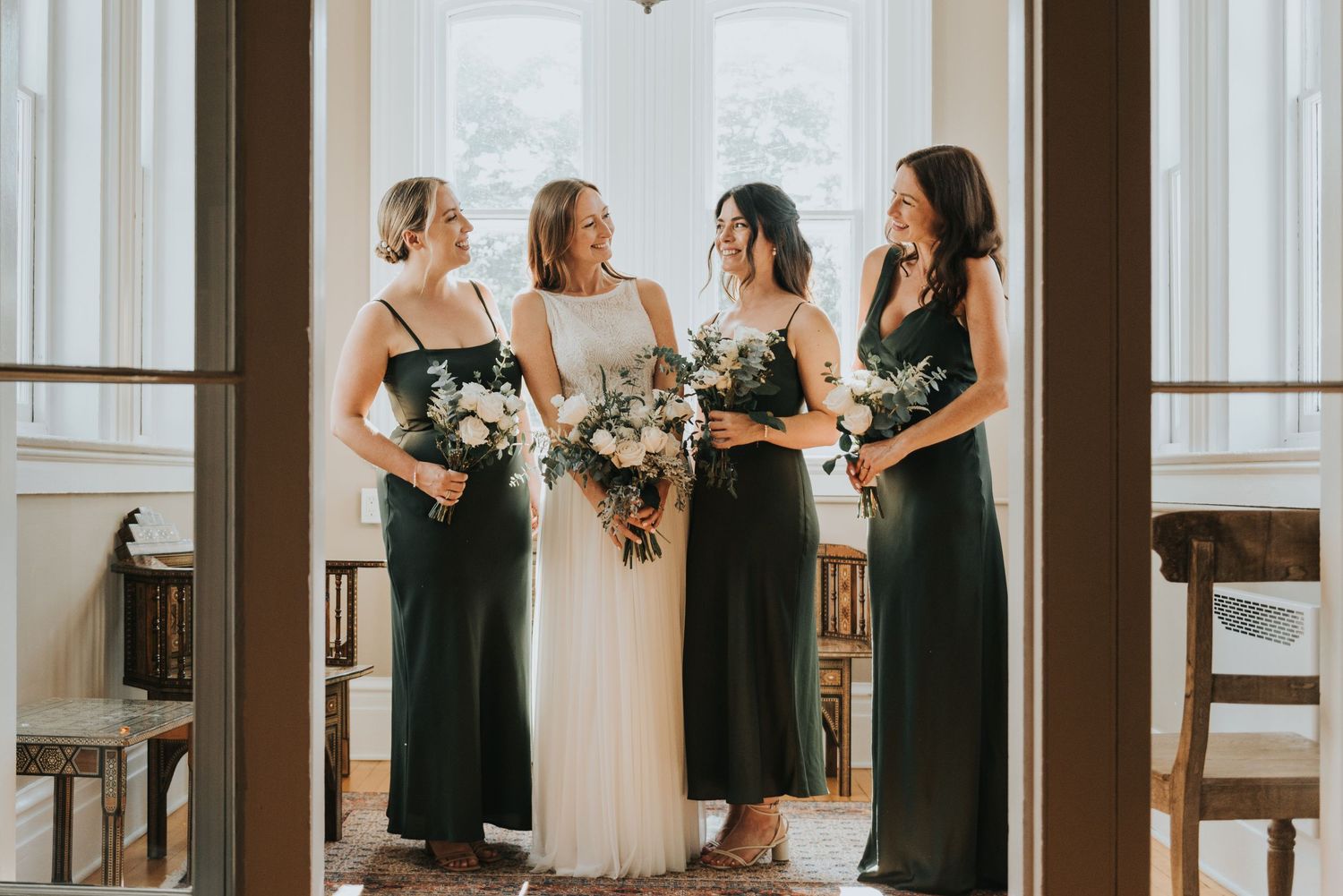 A bridal party in formal attire poses near large windows, with bridesmaids wearing elegant dark dresses holding bouquets.