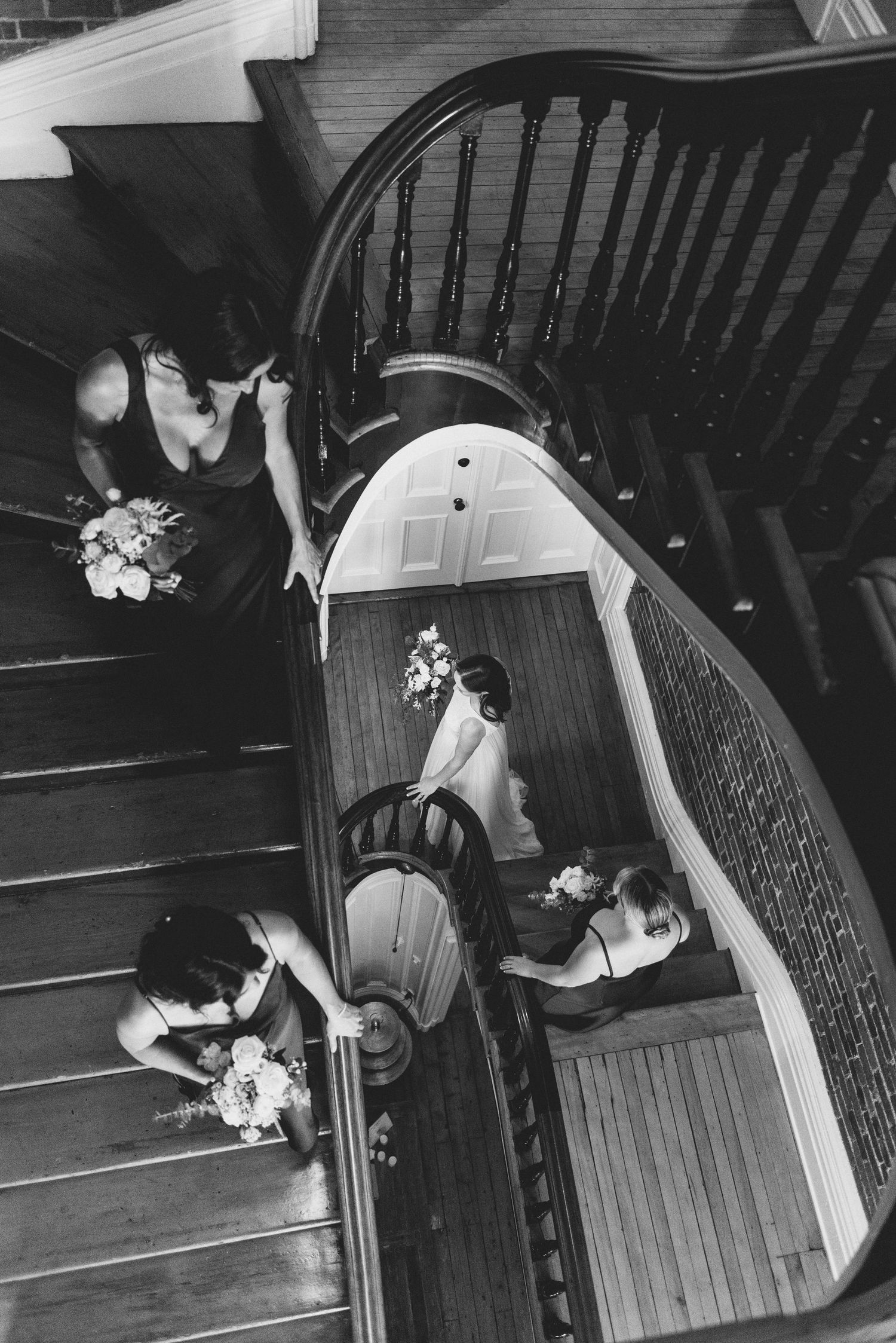 Black and white photo of a wedding party arranged on a curved staircase, shot from above showing floral bouquets.