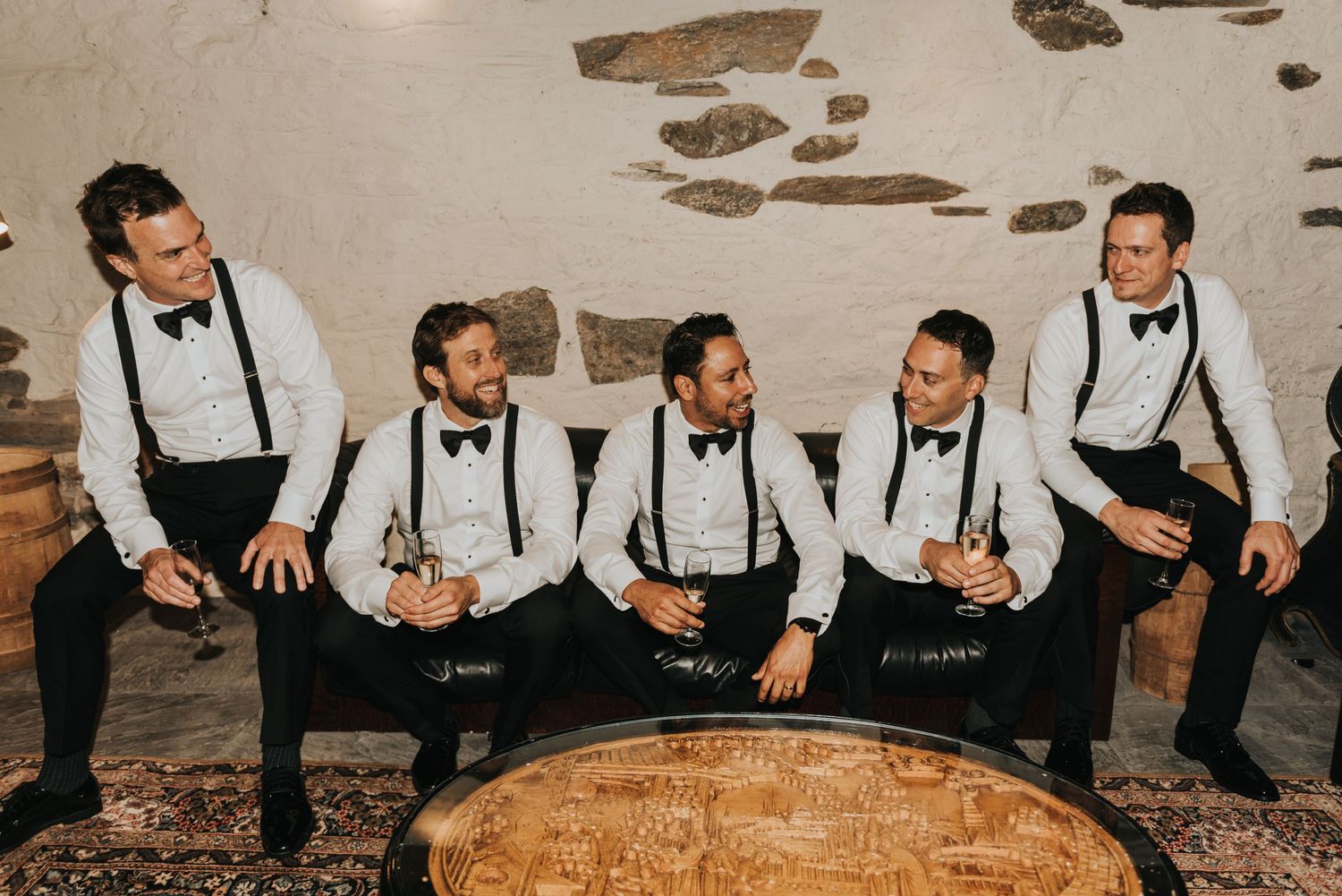 Groomsmen in matching black suspenders and bowties sitting casually on a rustic bench against a weathered wall.