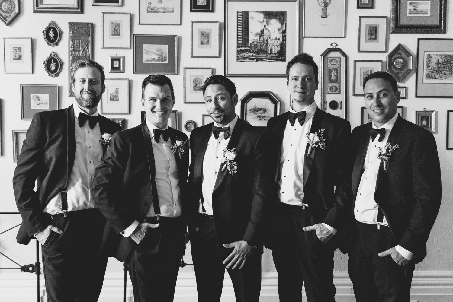 Groomsmen in formal black tuxedos posing against wall decorated with vintage framed photos.