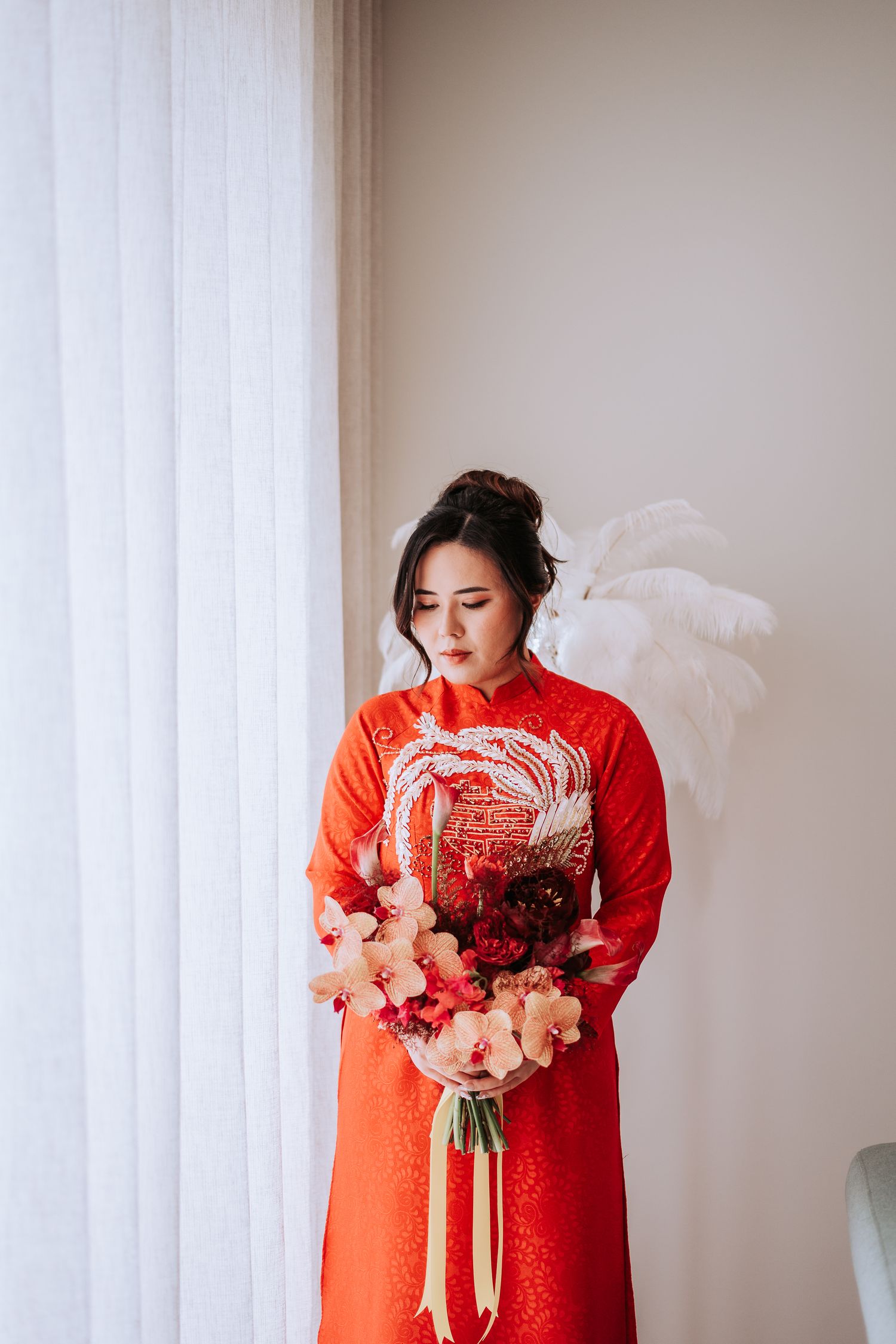 A figure in a traditional red Vietnamese ao dai dress holds a pink and red floral bouquet by a window.