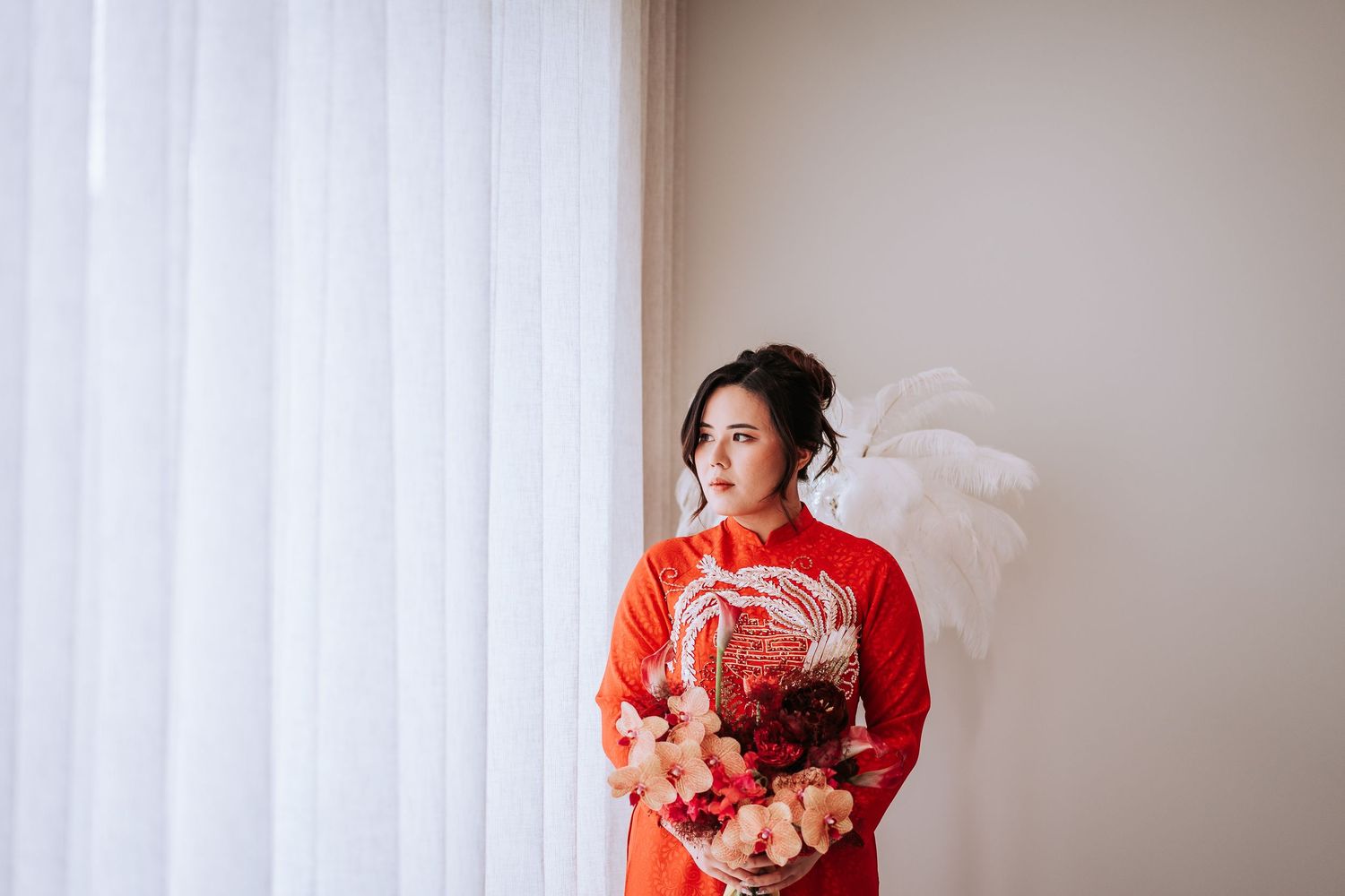 Woman in red Chinese traditional qipao dress holding bouquet stands by window with soft curtains.