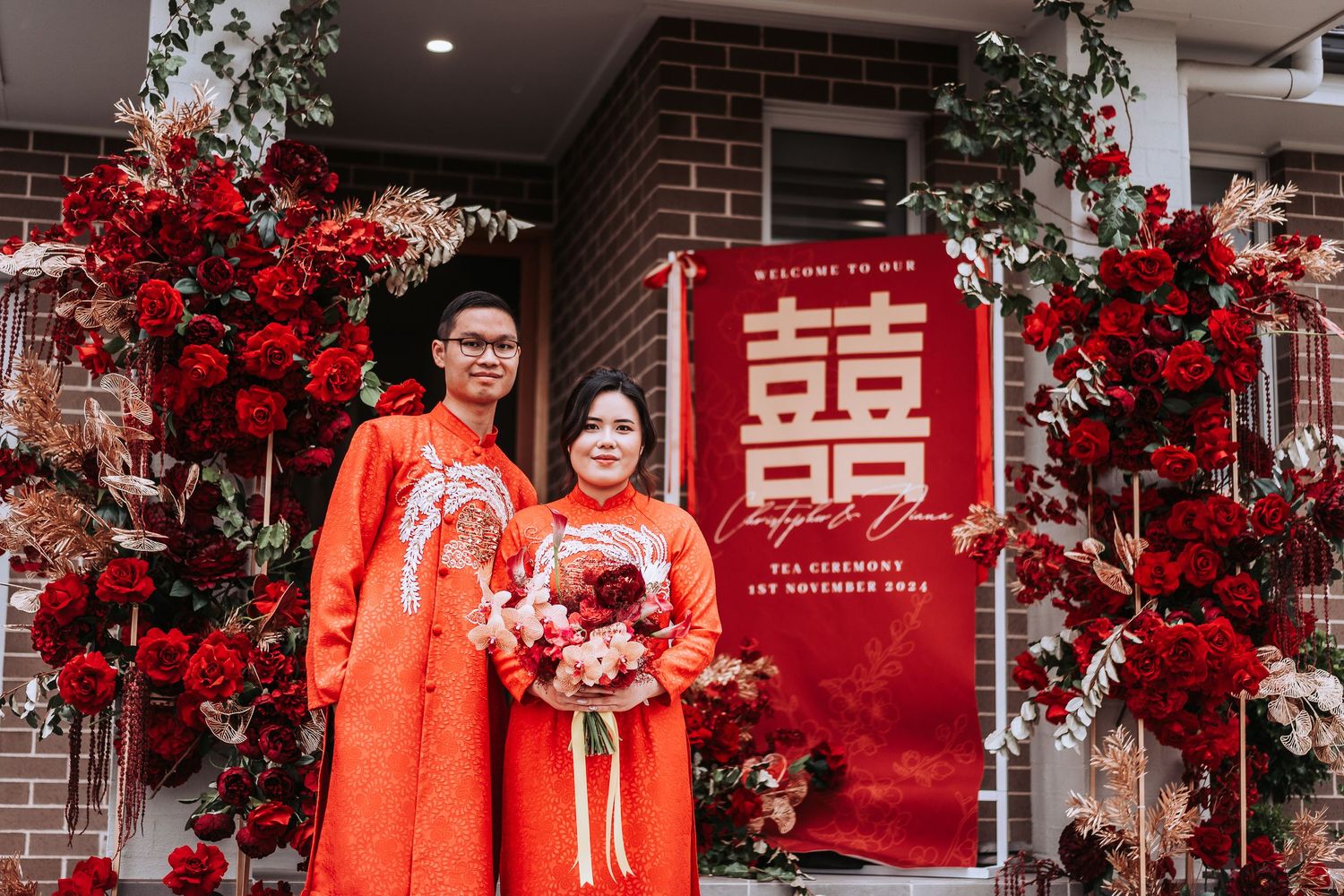 Couple in red traditional outfits pose by decorated entrance with double happiness symbol and red flowers.