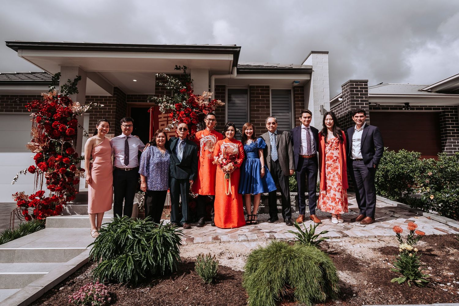 Group photo of wedding party members in formal attire standing outside a modern house with red floral decor.