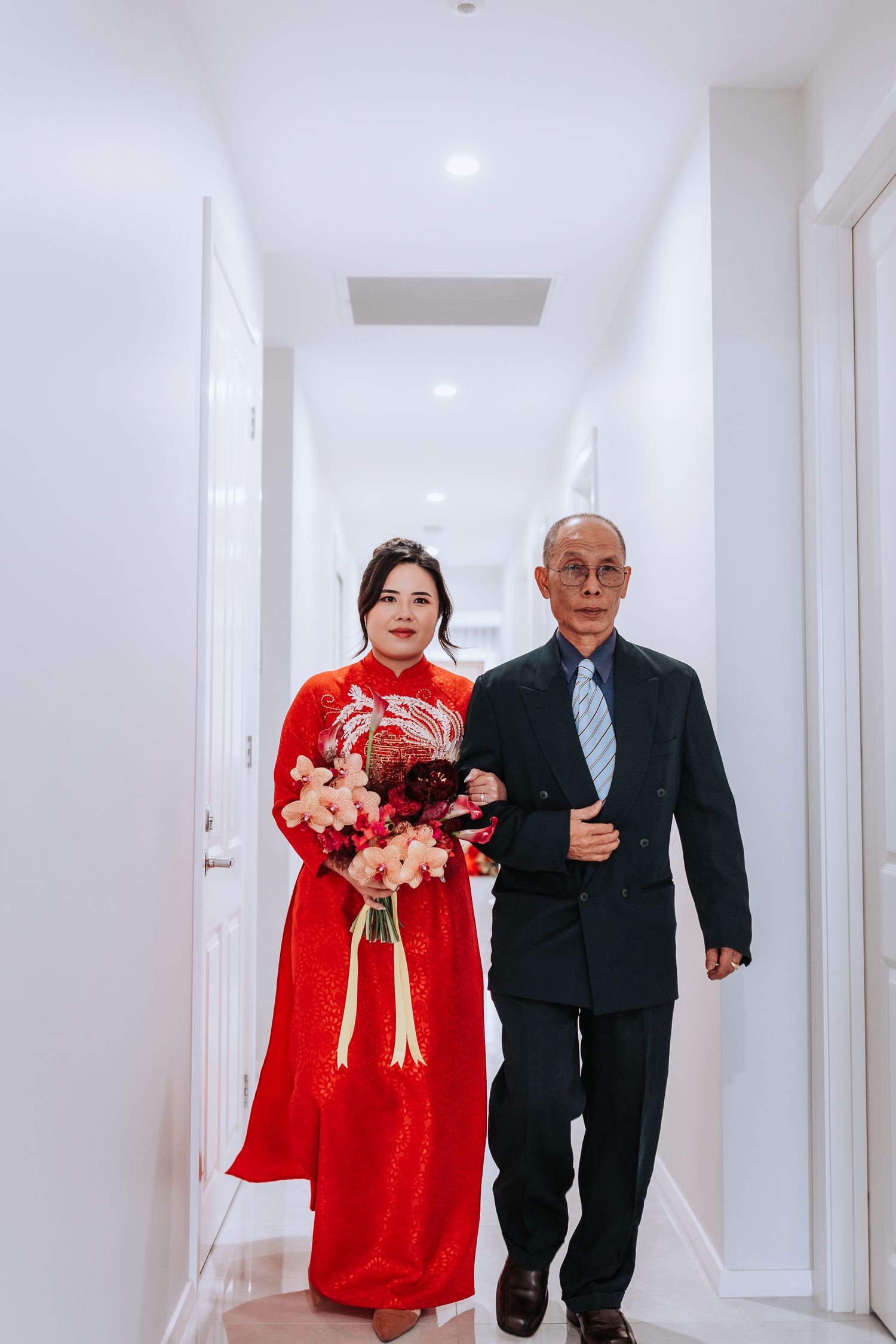 Two people walking down a white hallway, with one wearing a traditional red dress holding flowers and another in a suit.