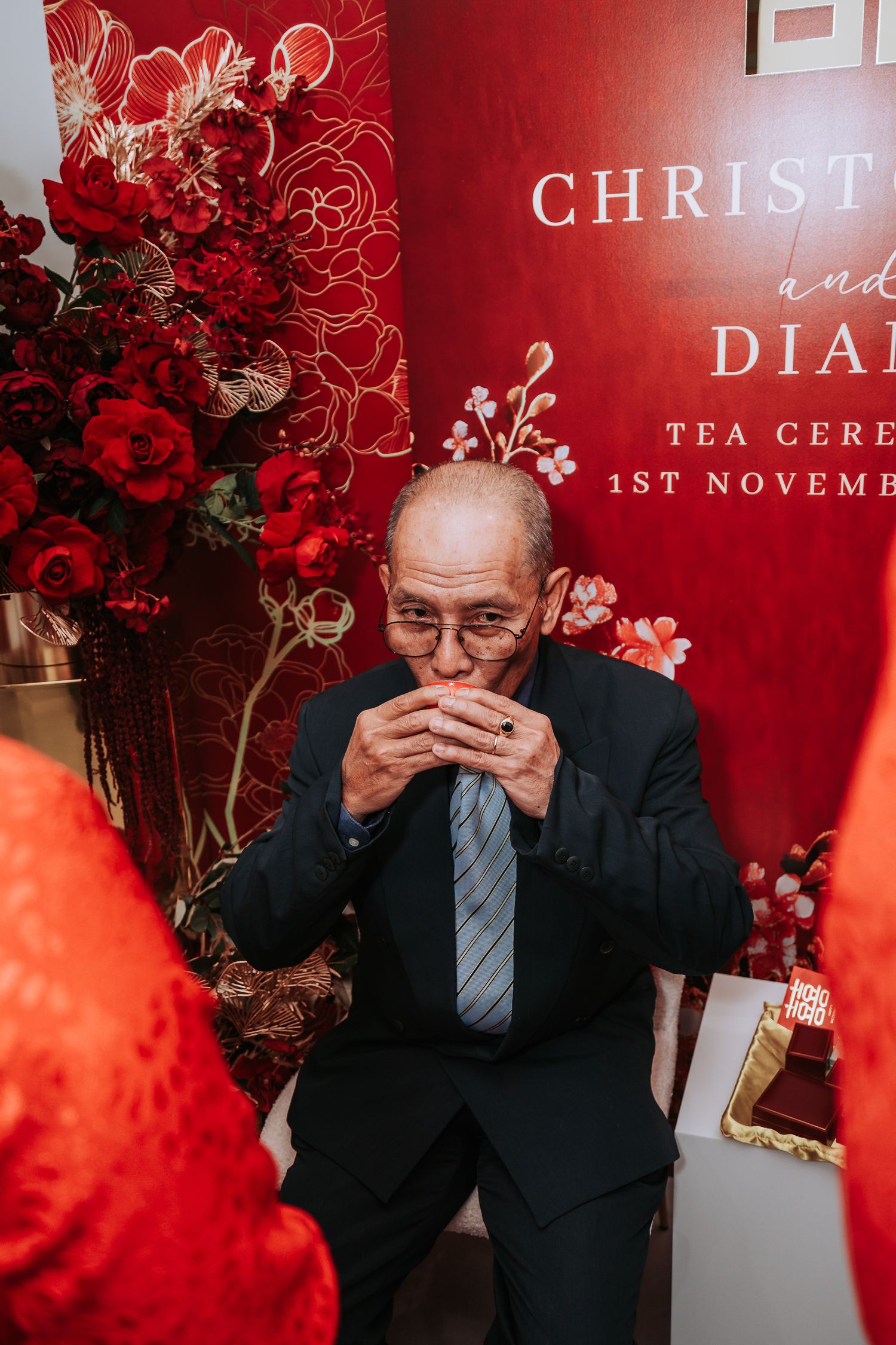 A person in a dark suit and tie sips a drink at a formal event with red floral decor and signage in the background.