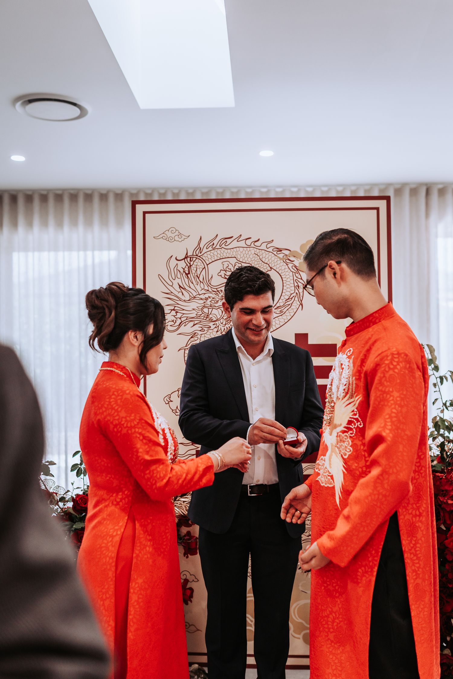 Wedding ceremony with couple in red traditional Chinese attire exchanging rings with officiant in black suit.