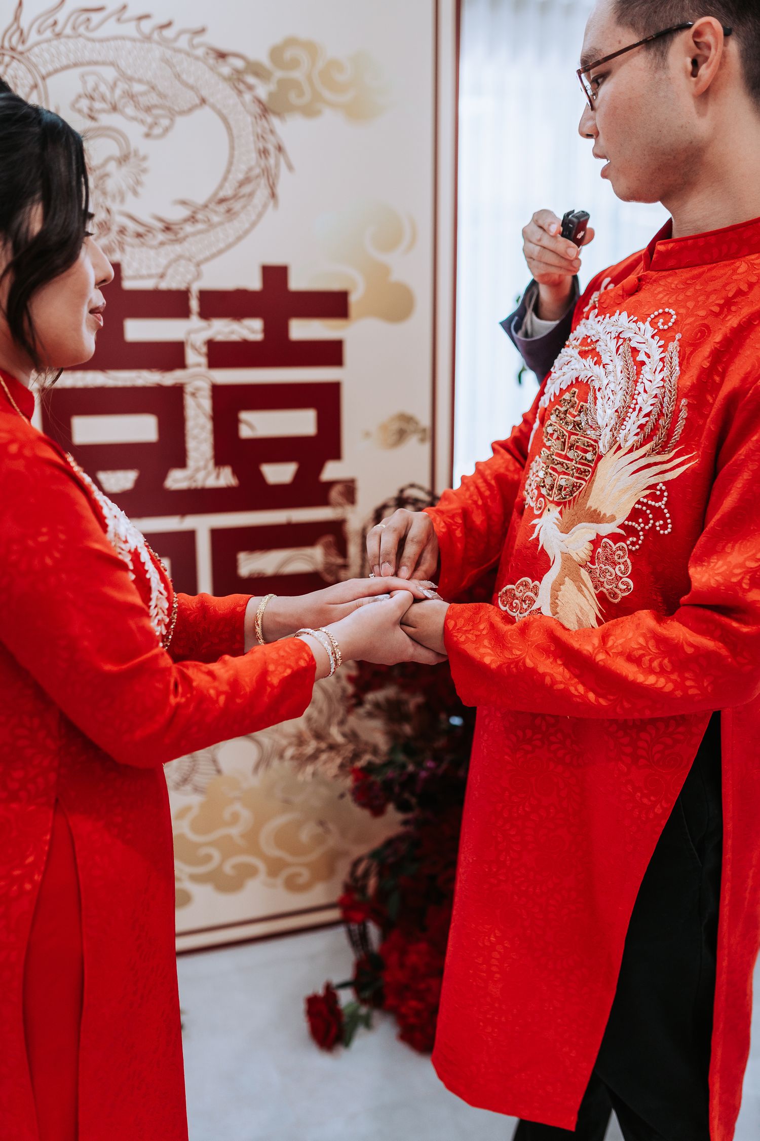 Close up of couple in red traditional Chinese wedding attire exchanging rings with double happiness symbol in background.
