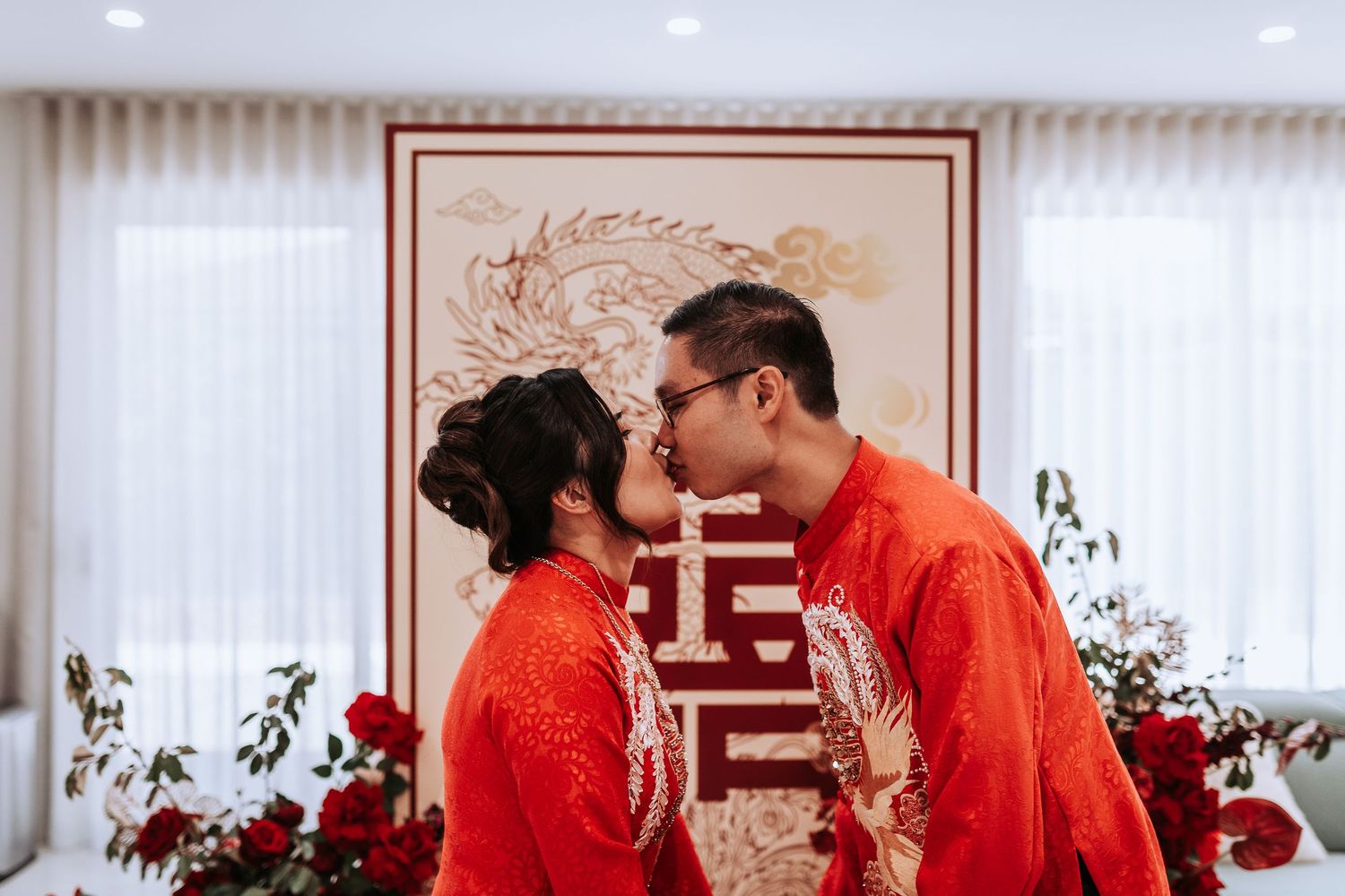 Traditional Chinese wedding ceremony with couple in red qipao and changshan attire kissing in front of decorative backdrop.
