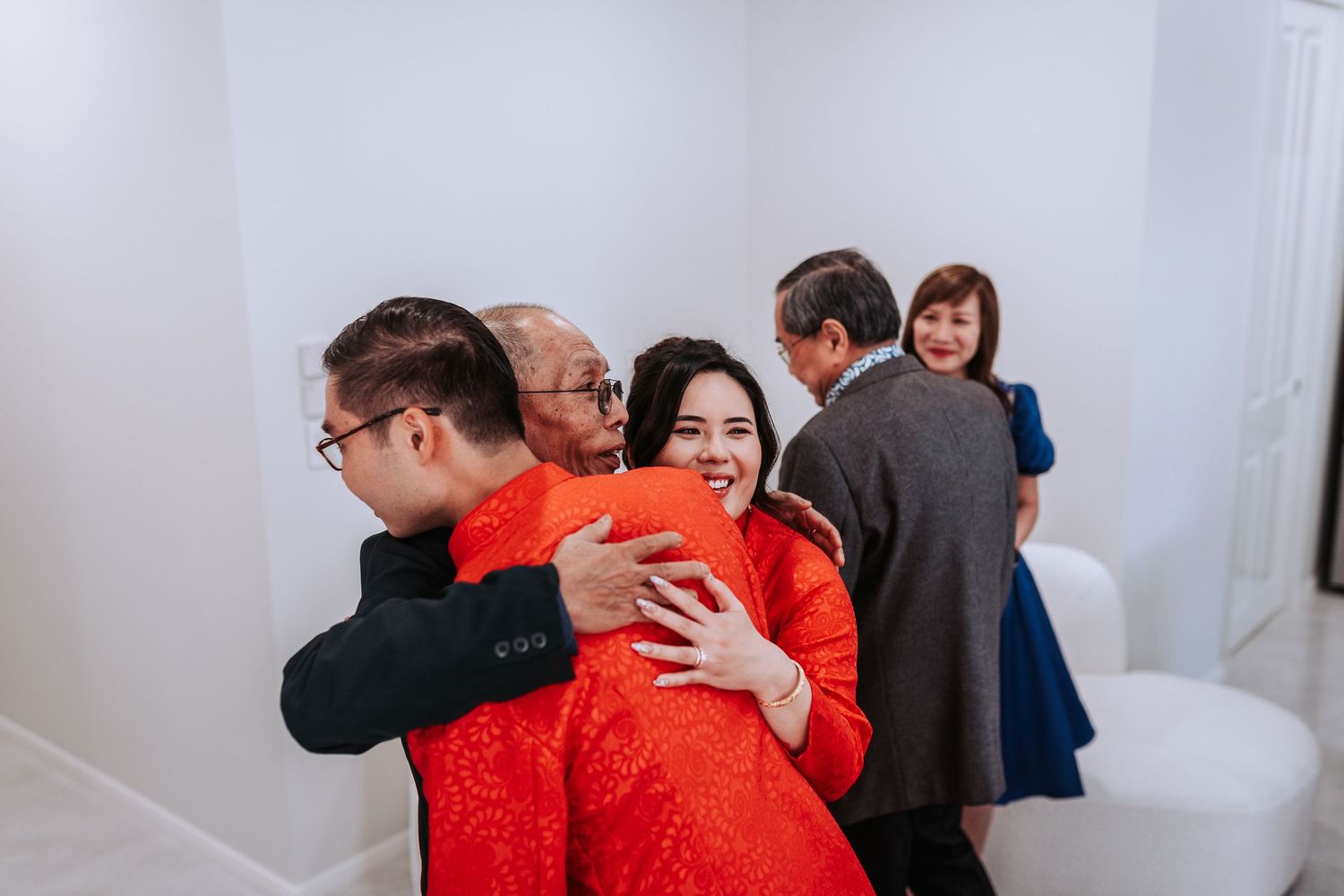 People embracing at an indoor event, with one person wearing a vibrant red outfit against a white wall background.