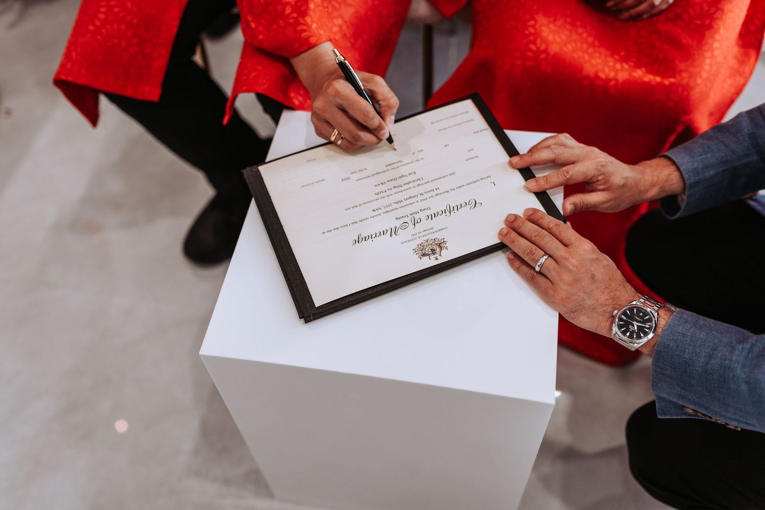 Hands signing a marriage certificate or legal document on a white surface with someone wearing a bright red outfit.