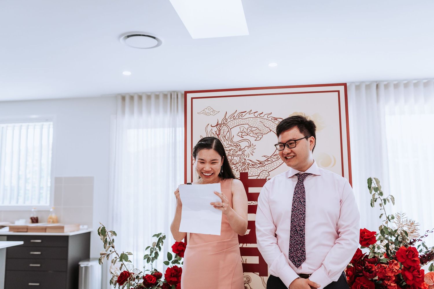 Two people in business attire smile while standing together by red floral arrangements and decorative wall art.