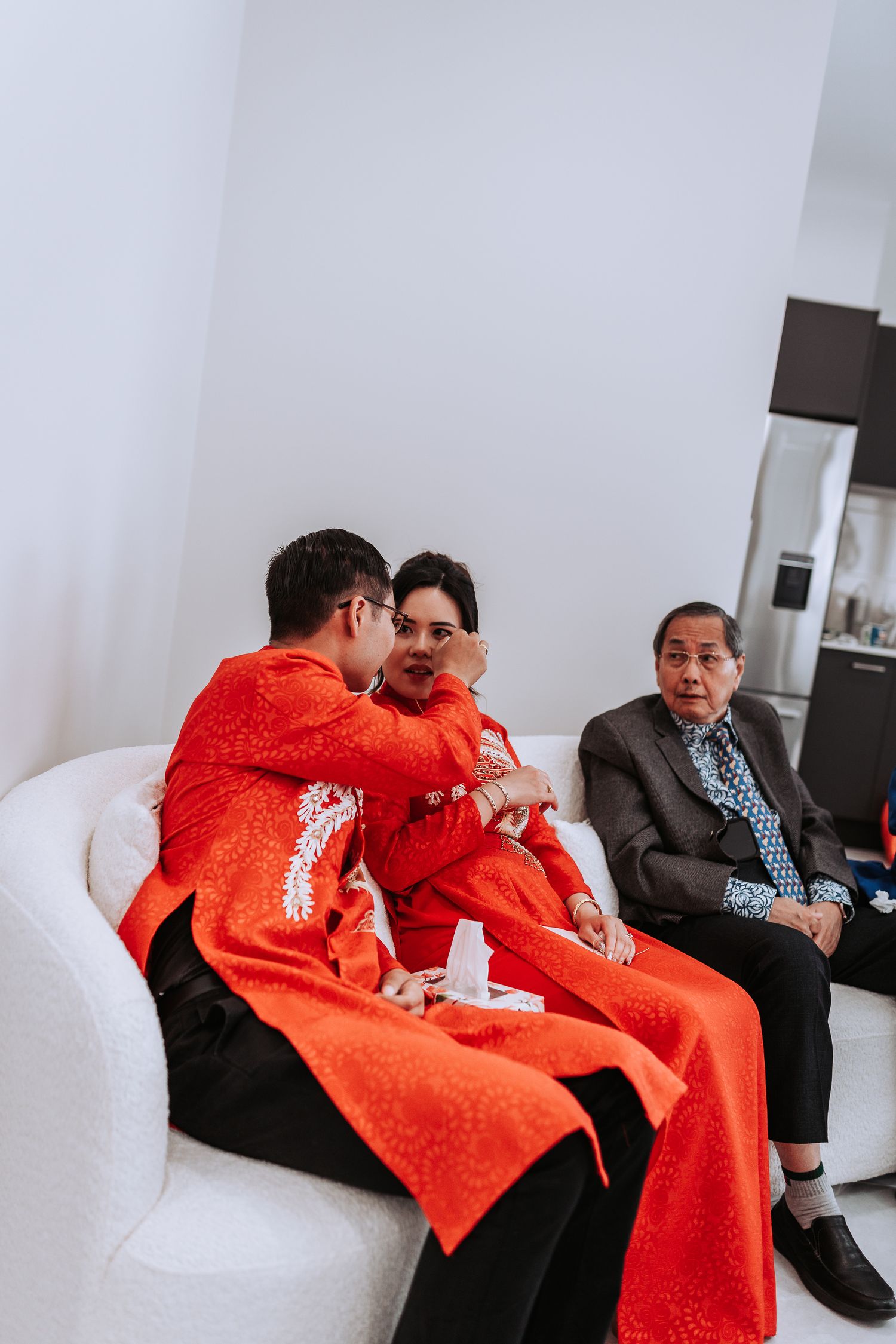 A couple in traditional bright red Chinese wedding attire sits together on a white couch while a guest looks on.