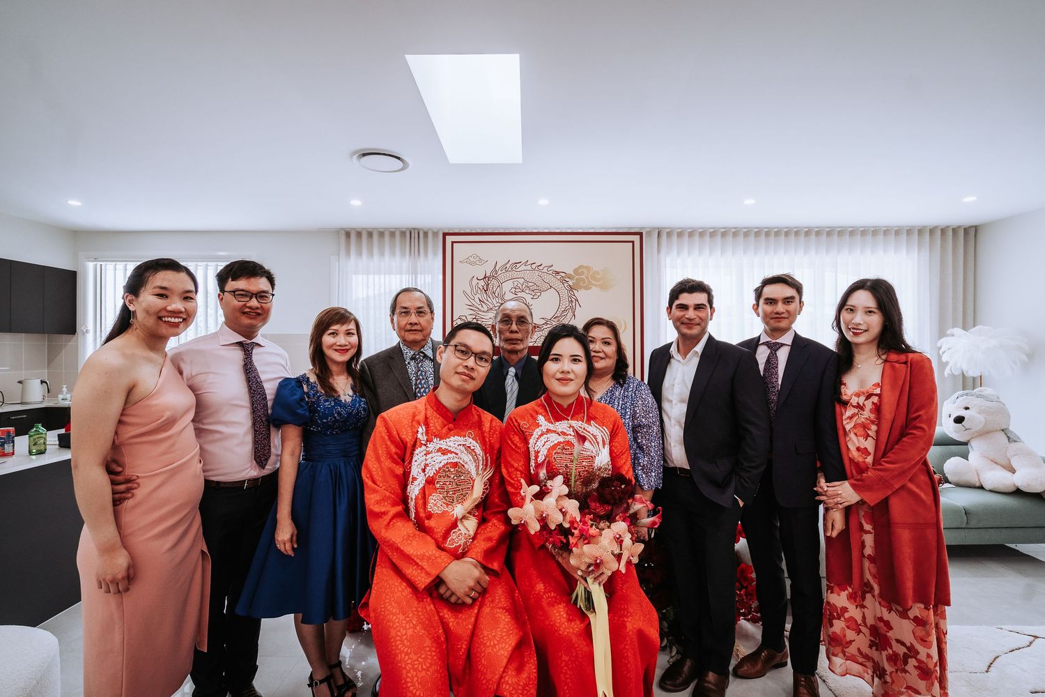 Wedding party gathering indoors with people wearing mix of traditional red Chinese outfits and formal Western attire.