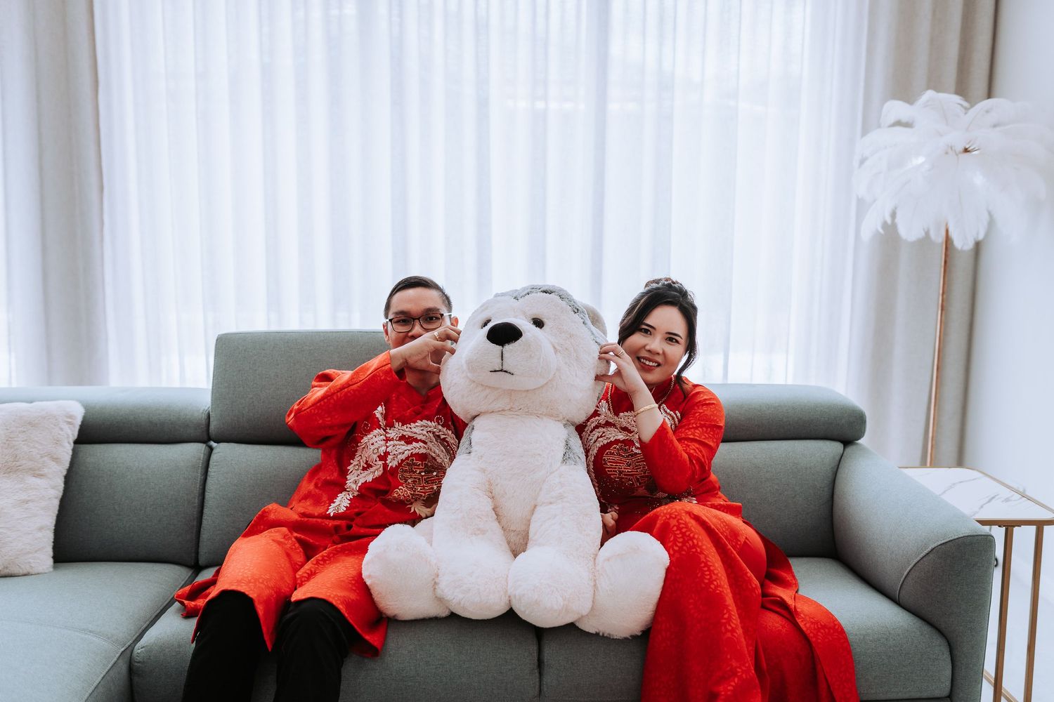 A couple in matching red outfits pose with a large white teddy bear on a grey couch.