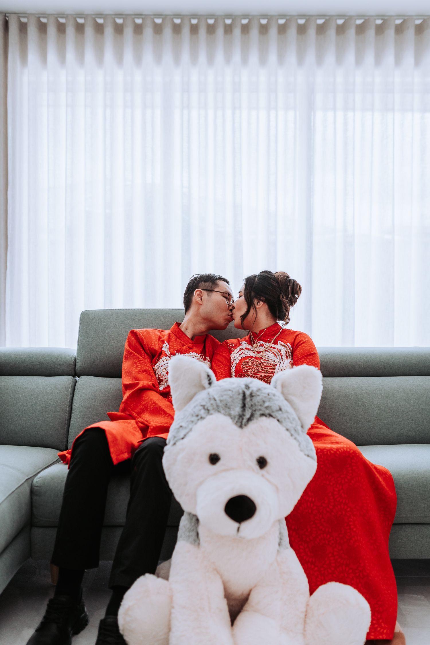 A romantic couple in matching red traditional outfits sits on a gray couch with a large white plush polar bear.