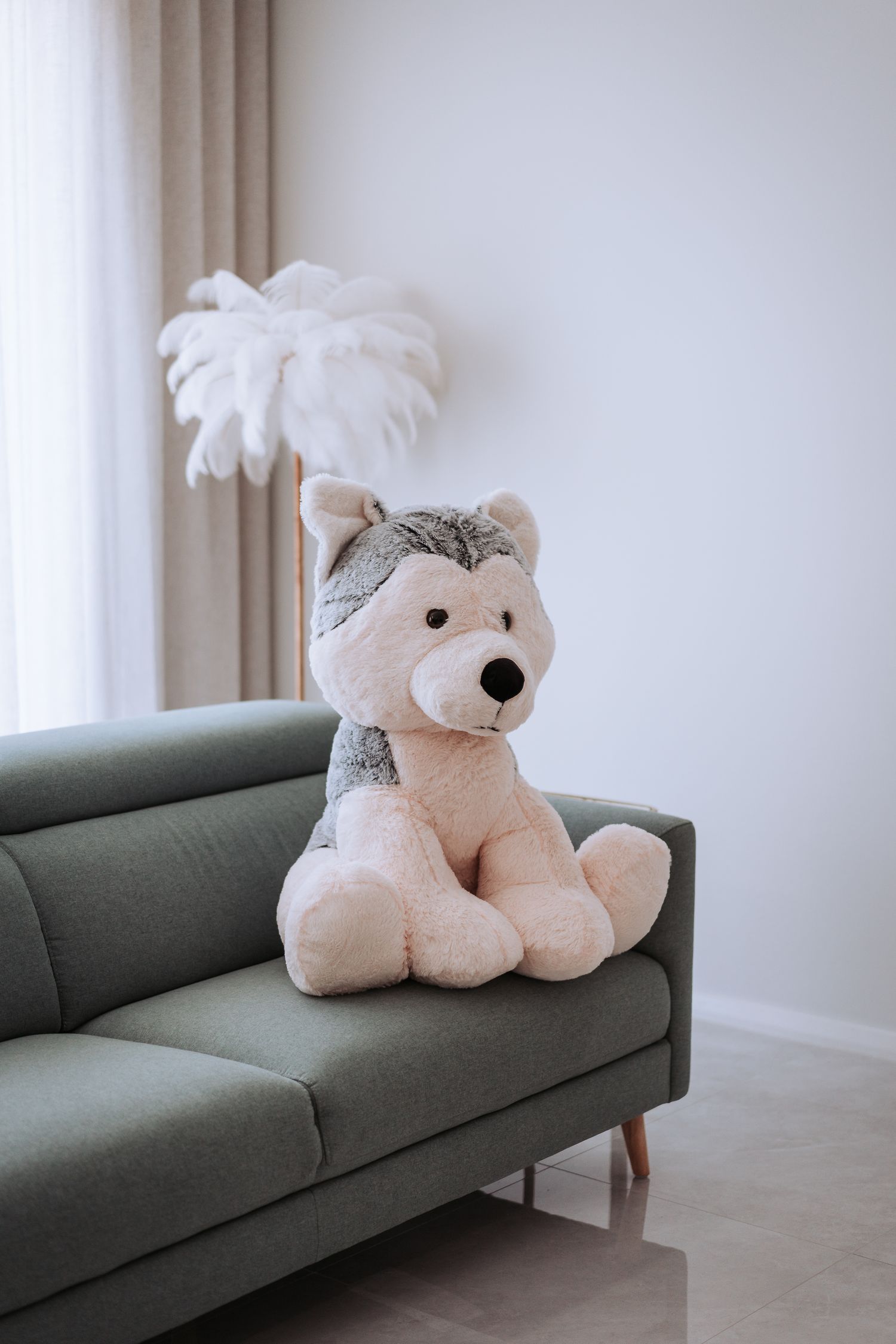 A plush gray teddy bear sits on a sage green couch with a white feather decoration in the background.