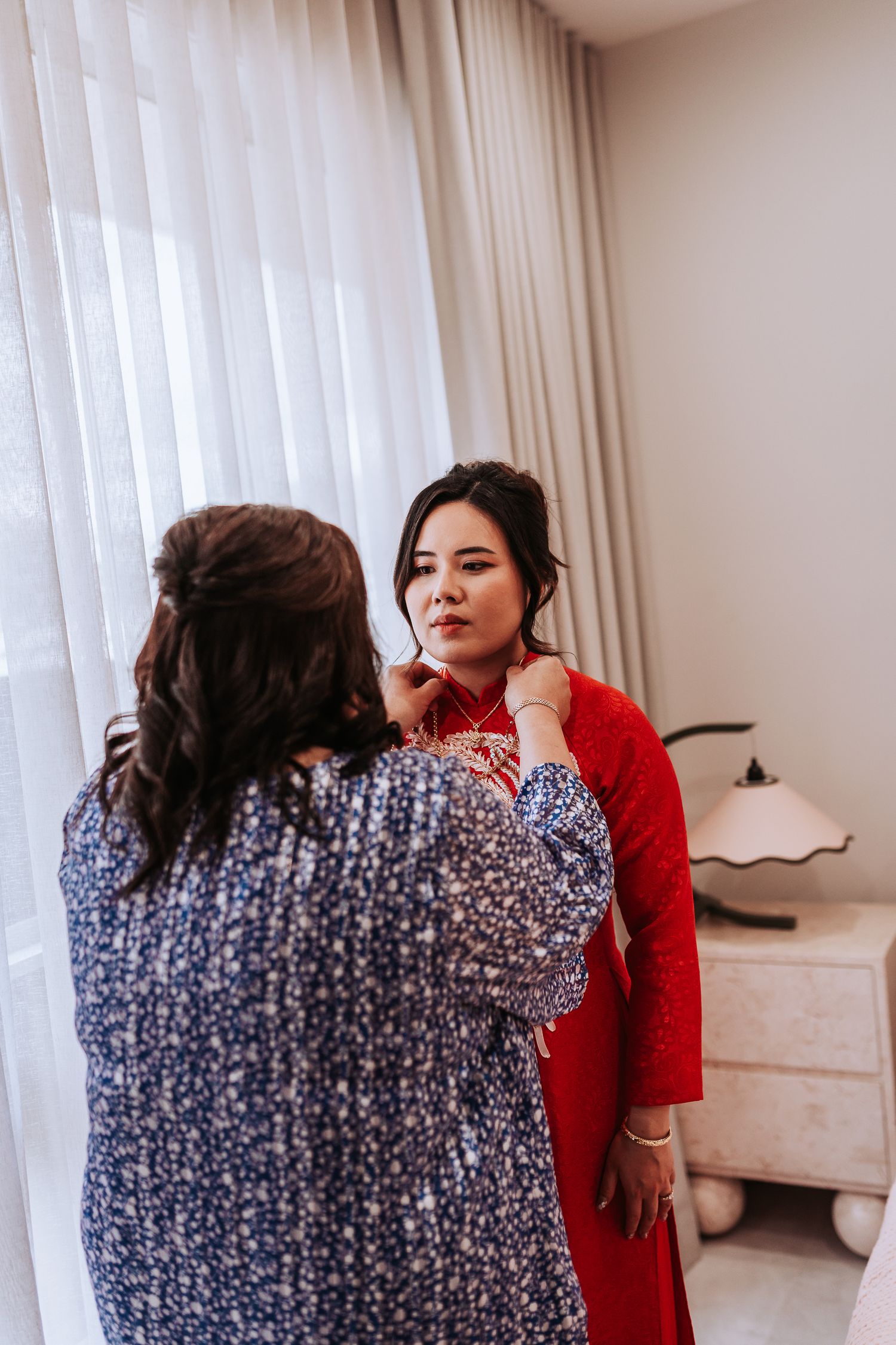 A woman in a red traditional Chinese qipao dress gets makeup applied while standing near white curtains in a bedroom.