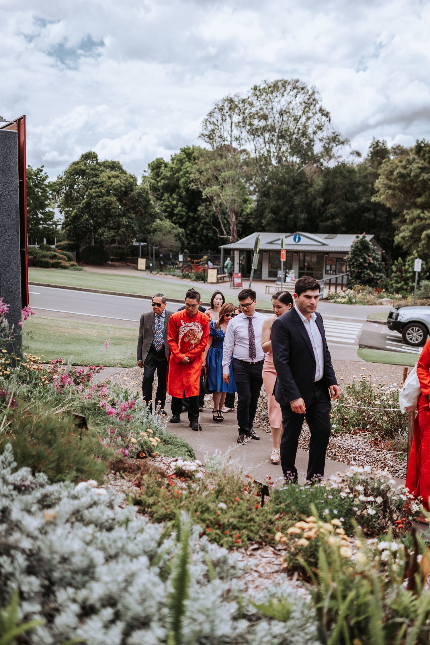 A group of formally dressed people walking along a garden path with blooming flowers and trees in the background.