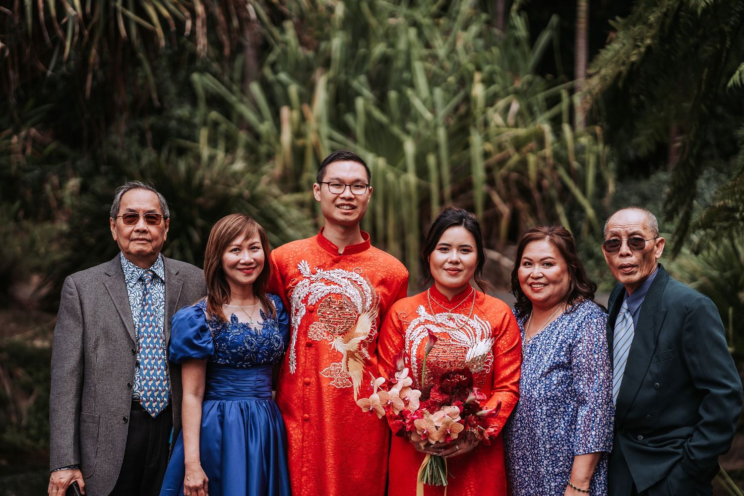 A group photographed at a traditional wedding ceremony wearing red and blue formal attire with tropical foliage backdrop.
