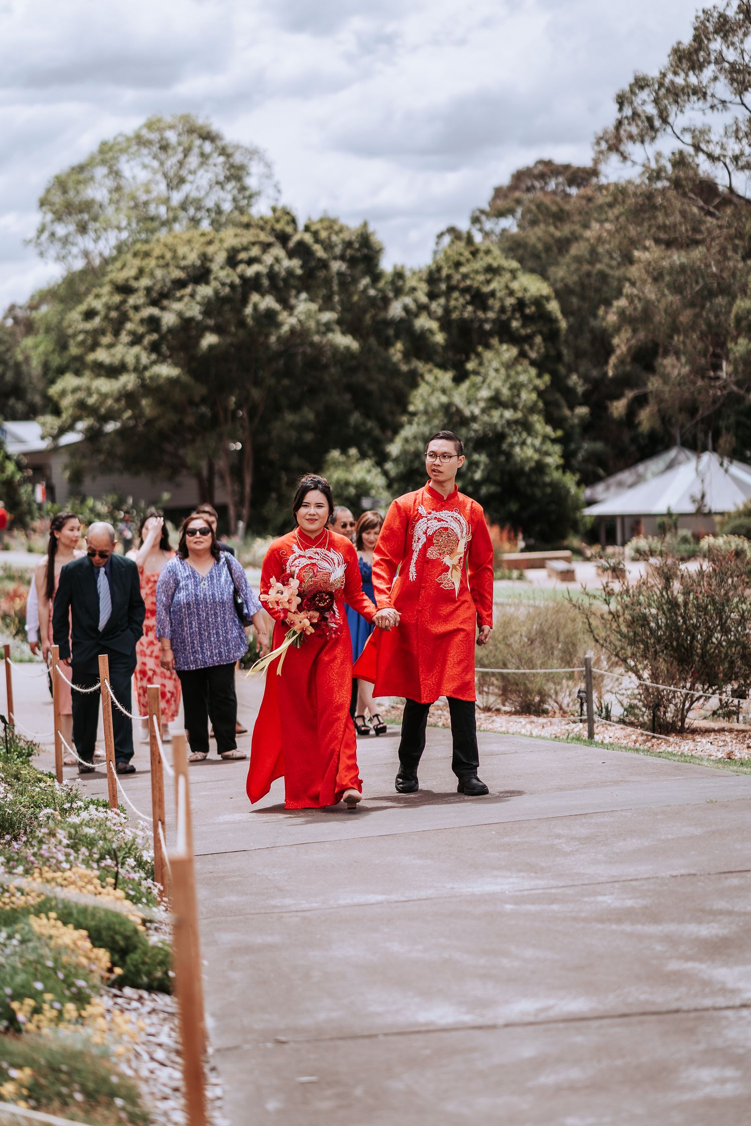 Wedding couple in traditional red Chinese attire walk along garden path with guests during traditional tea ceremony.