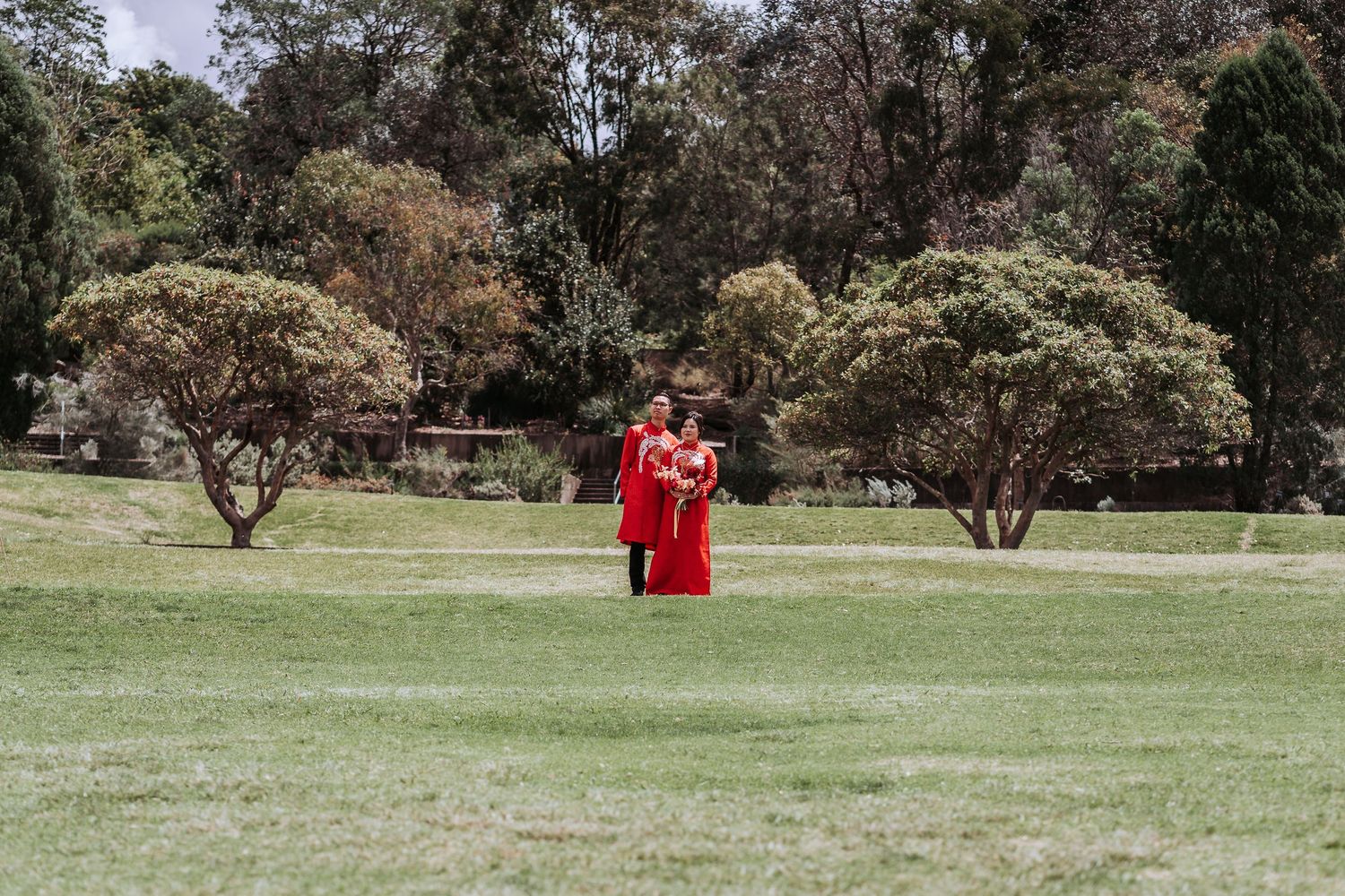 Two people in bright red attire stand together in a serene park setting surrounded by trees and green grass.