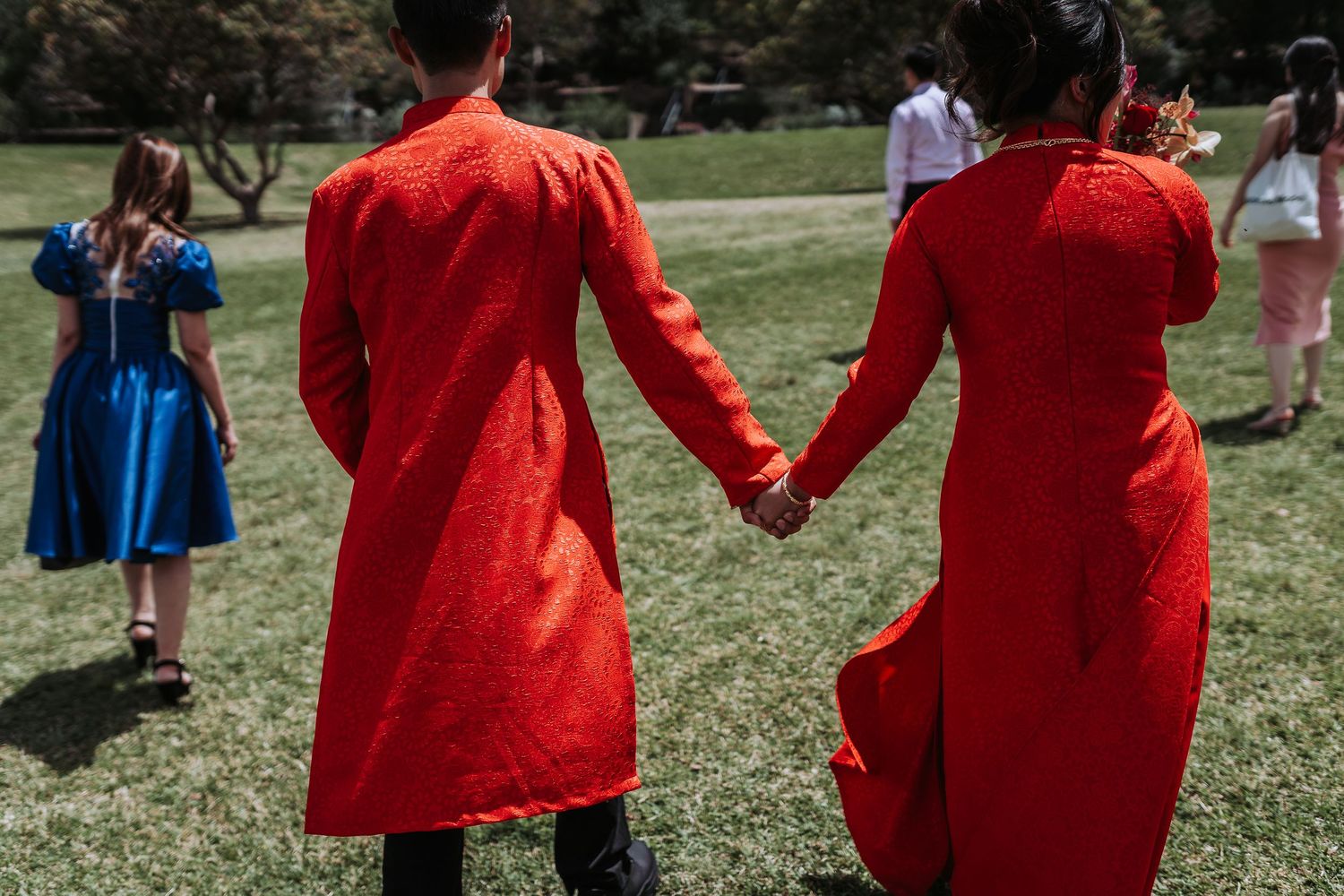 Two people in matching bright red traditional outfits hold hands on a grassy lawn during an outdoor gathering.