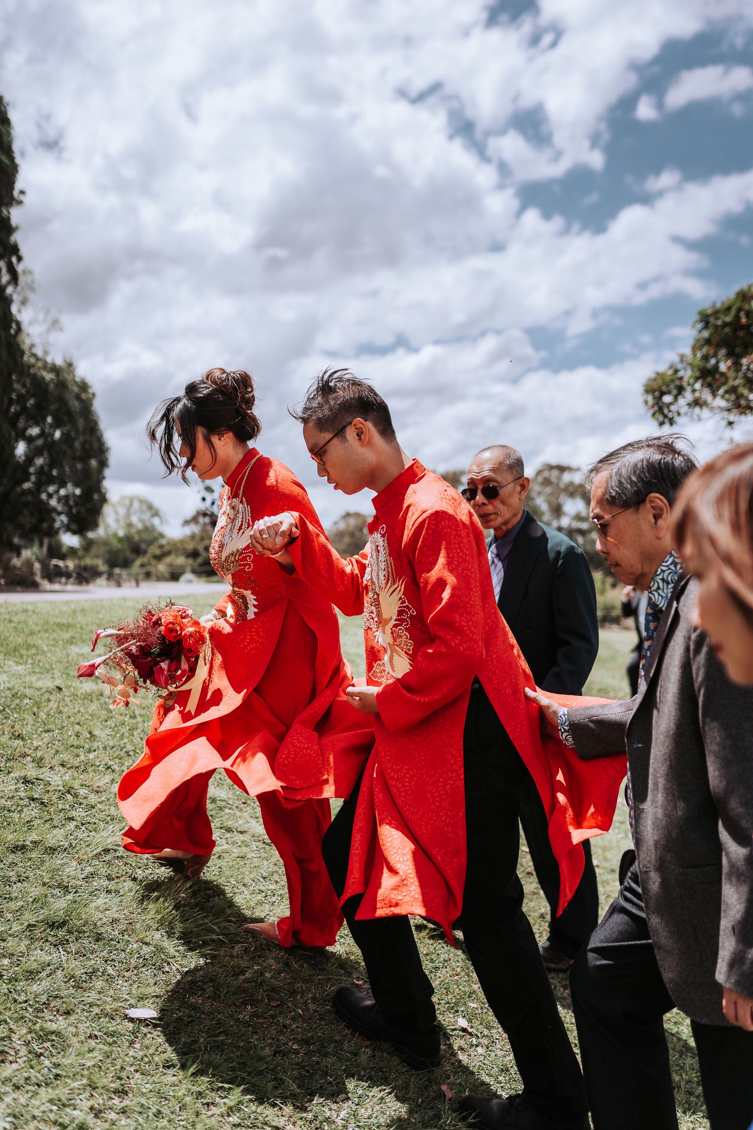 A group in bright red traditional attire performing a ceremonial walk outdoors on grass beneath a cloudy sky.