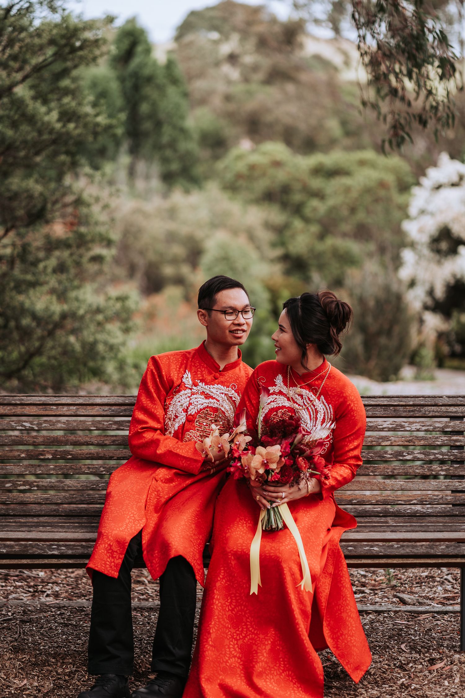 Couple in traditional red ao dai attire sitting together on wooden bench in garden setting with floral bouquet.