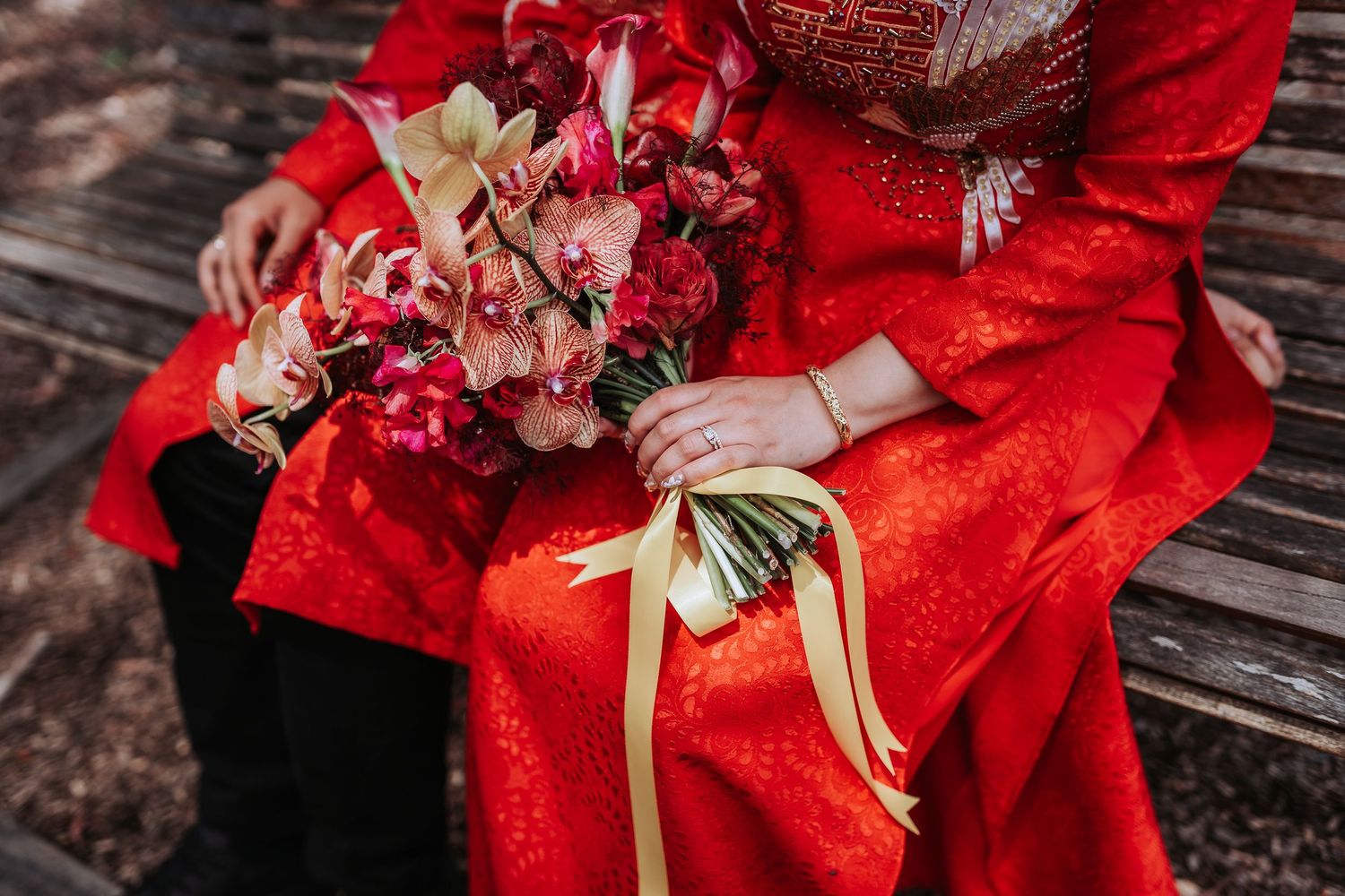 Person wearing a vibrant red dress holds an elegant bouquet of pink and blush flowers with gold ribbon on wooden bench.