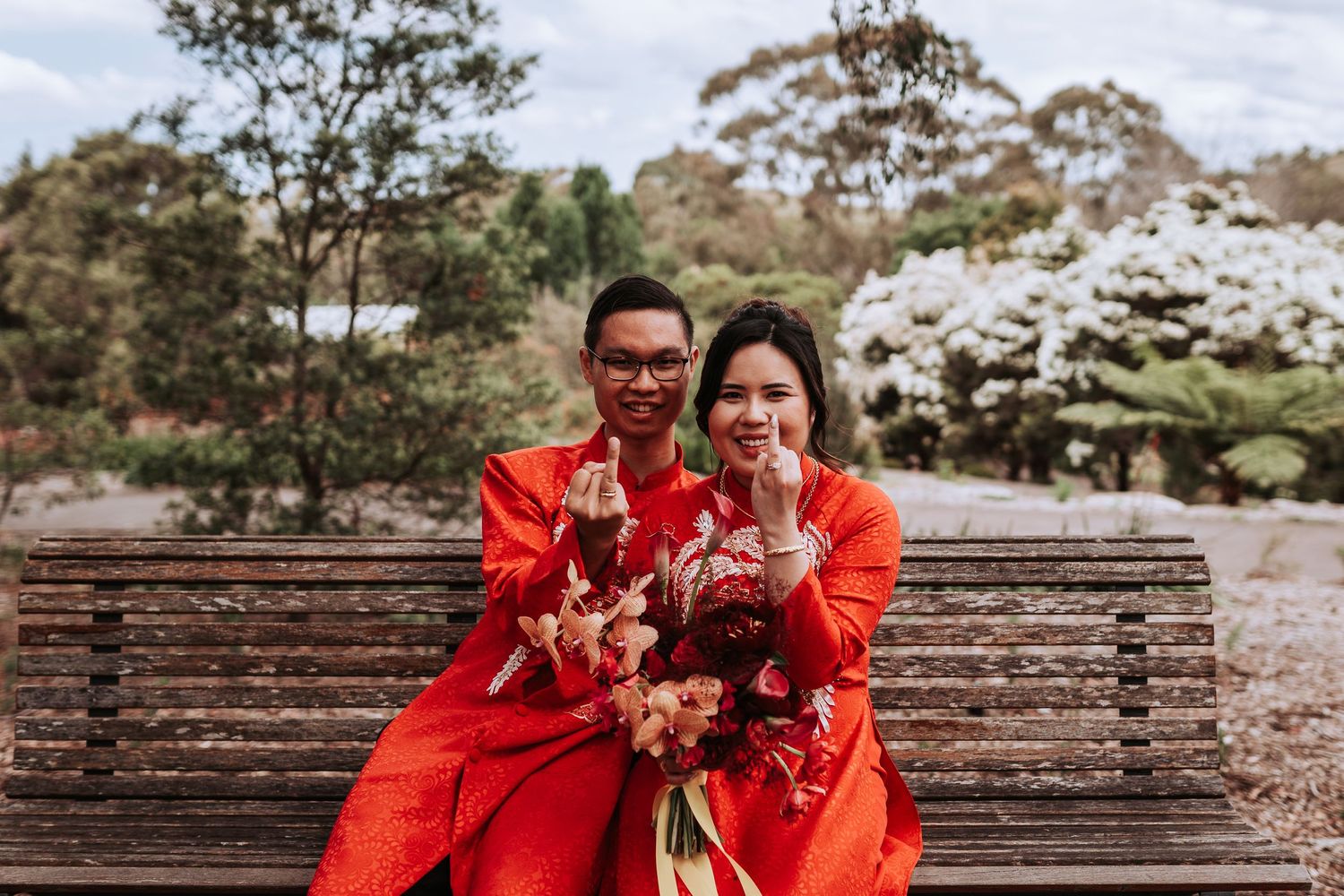Two people in matching red traditional outfits hold bouquets while sitting on wooden bench outside during spring.