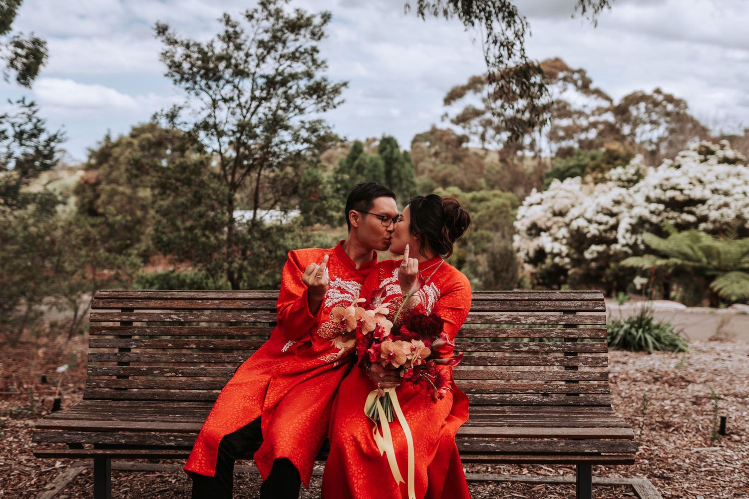 Couple in traditional red wedding attire sitting together on wooden bench in garden scenery.