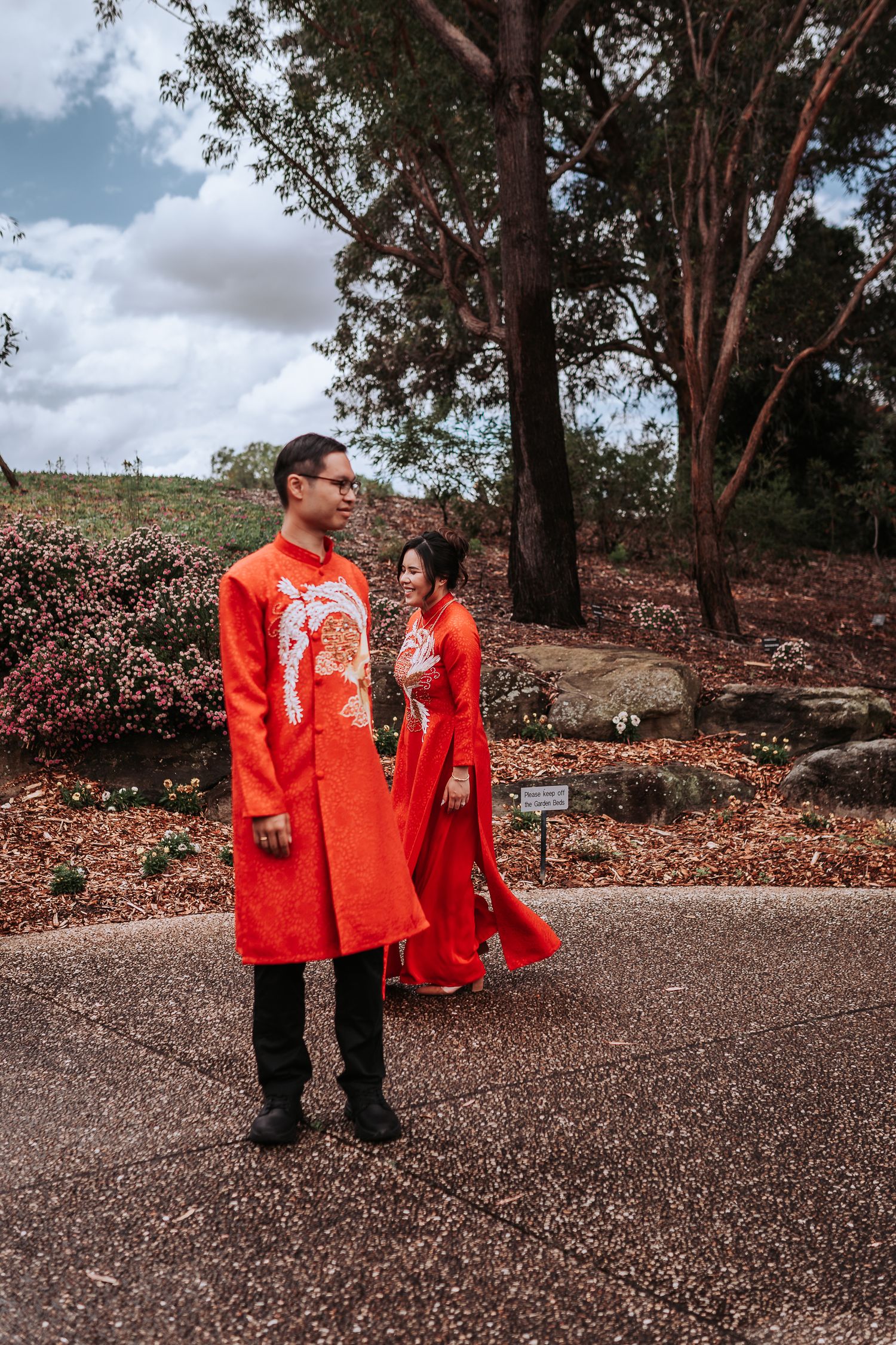 A couple in traditional red Chinese wedding attire stands together on a path beneath tall trees.