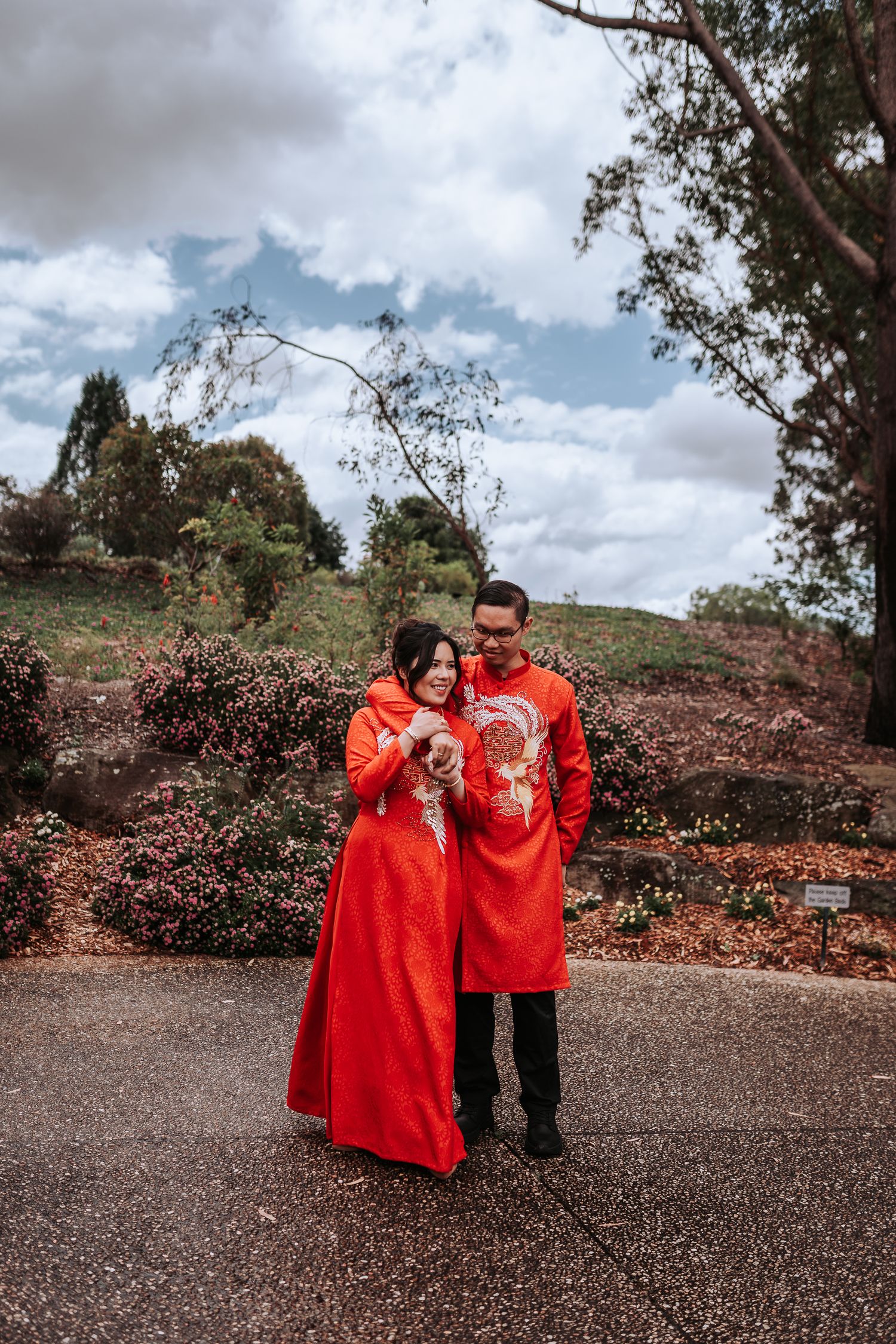 A couple wearing traditional red Chinese wedding attire stands together on a path surrounded by autumn foliage.