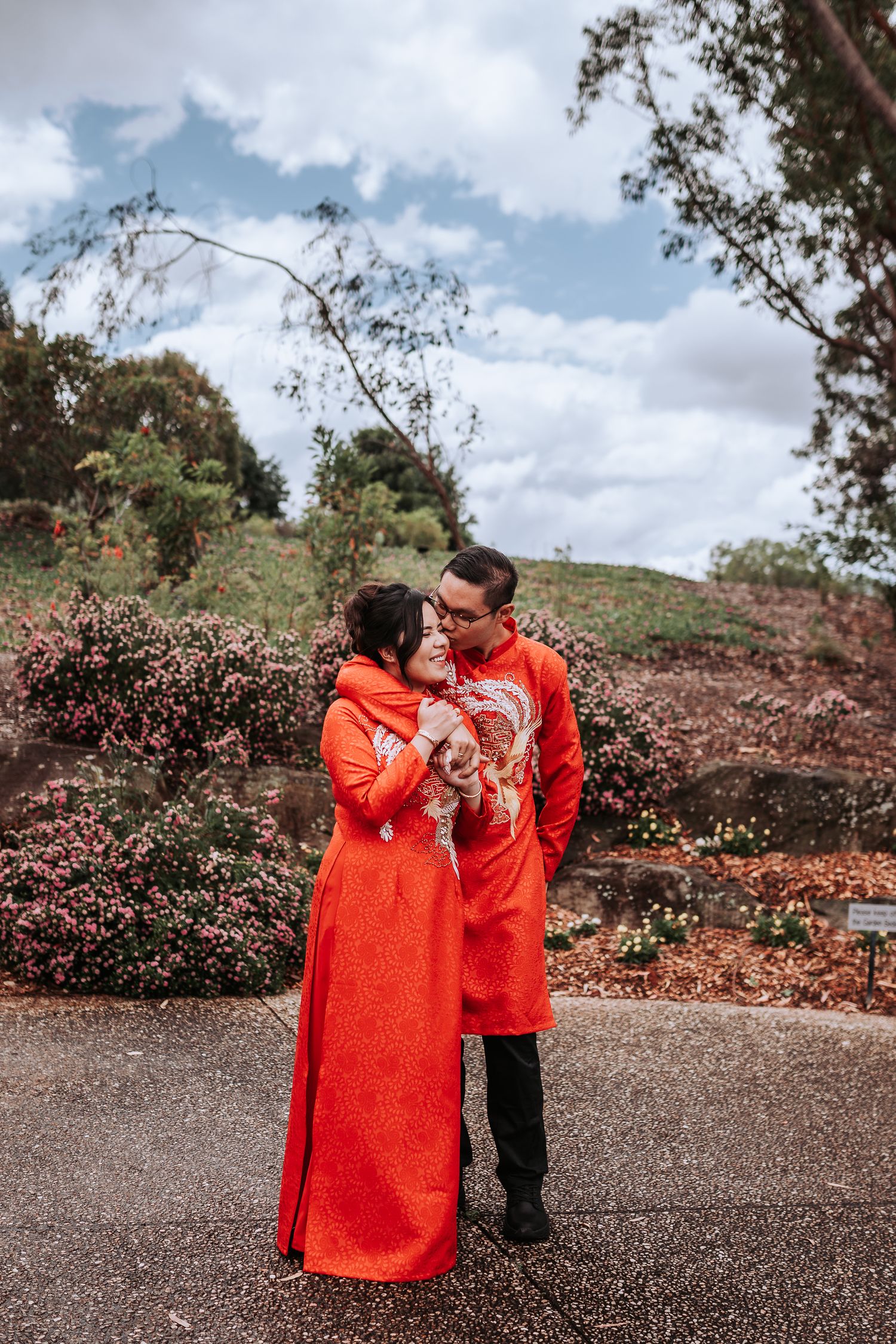 A couple in vibrant red traditional Chinese wedding attire embraces on a garden path under cloudy skies.