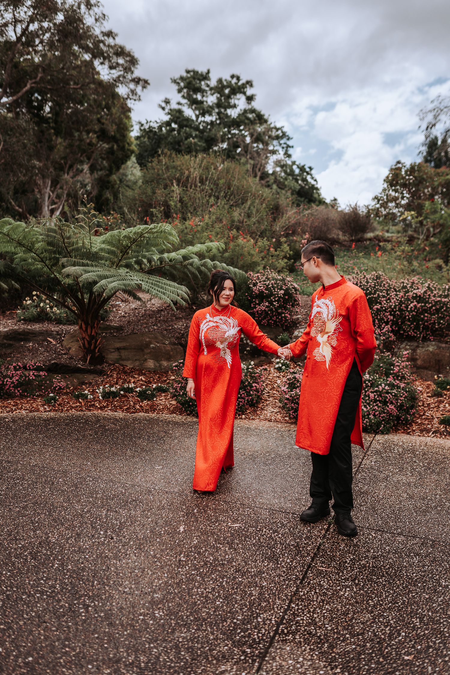 Couple in matching red traditional Asian formal attire holding hands on a garden pathway surrounded by ferns.