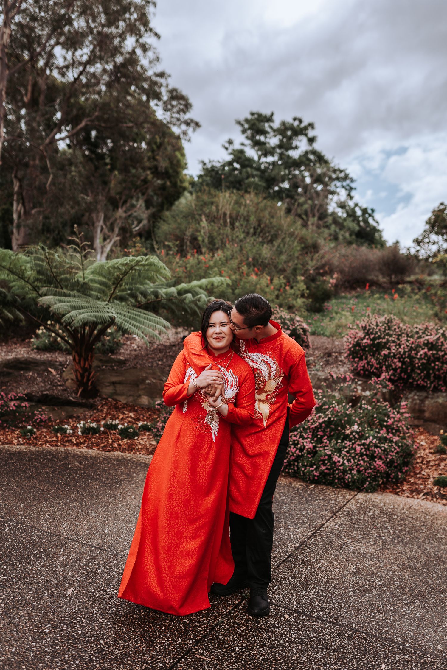 Two people wearing matching red traditional outfits share an intimate moment in a lush garden setting.